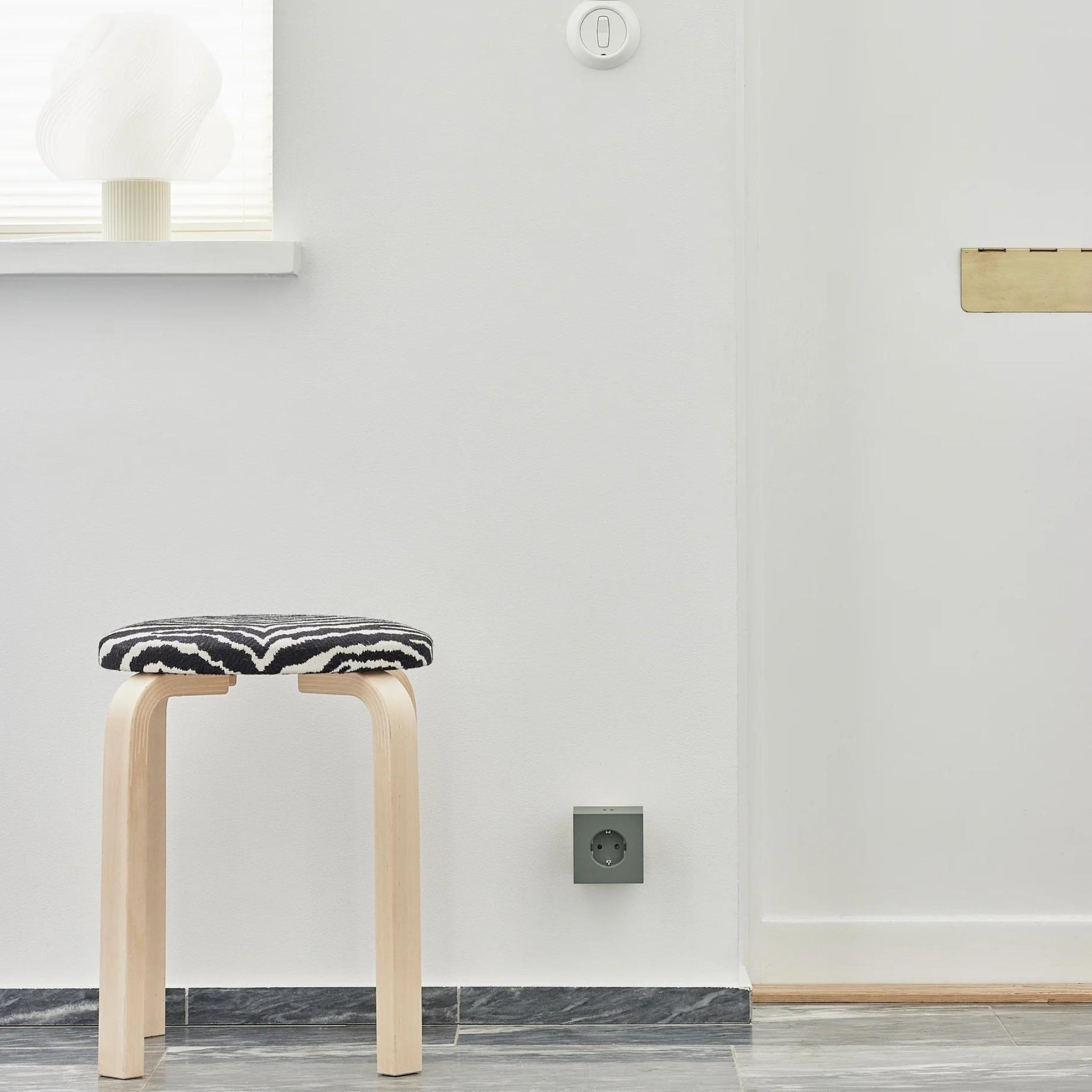 Een minimalistische kamer met een licht houten krukje, een zwart-wit kussen, een witte lamp op de vensterbank en een Avolt Square 2 USB-C Stekkerdoos Eik Groen van Avolt naast het stopcontact verbetert de neutrale ruimte met marmerdessin.