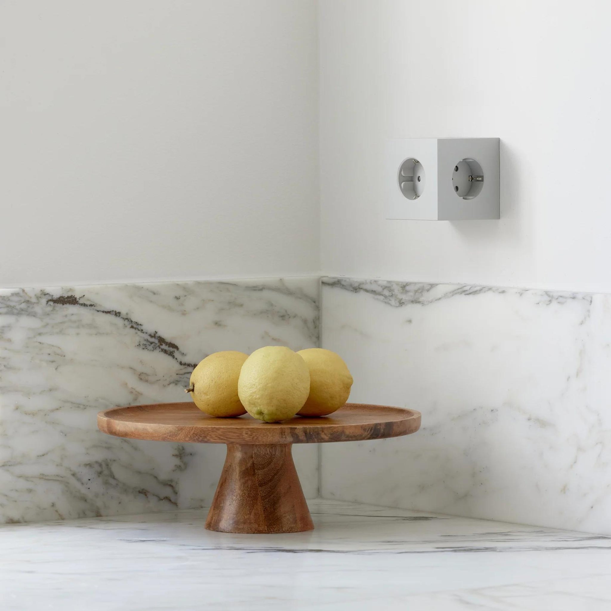 Een houten sokkel met drie citroenen staat op een wit marmeren aanrecht naast een Avolt Square 2 USB-C Stekkerdoos Gotland Grijs van Avolt, die naadloos opgaat in de witte muur en de bijpassende marmeren achterwand.