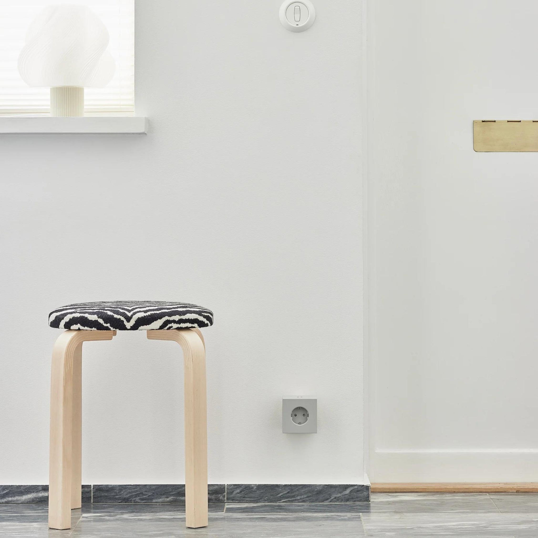 Een minimalistische kamer met een licht houten krukje met een zwart-witte zitting, een grijze vloer, een witte muur met een Avolt Square 2 USB-C Stekkerdoos Gotland Grijs van Avolt en een vensterbank met een wit decoratief object.