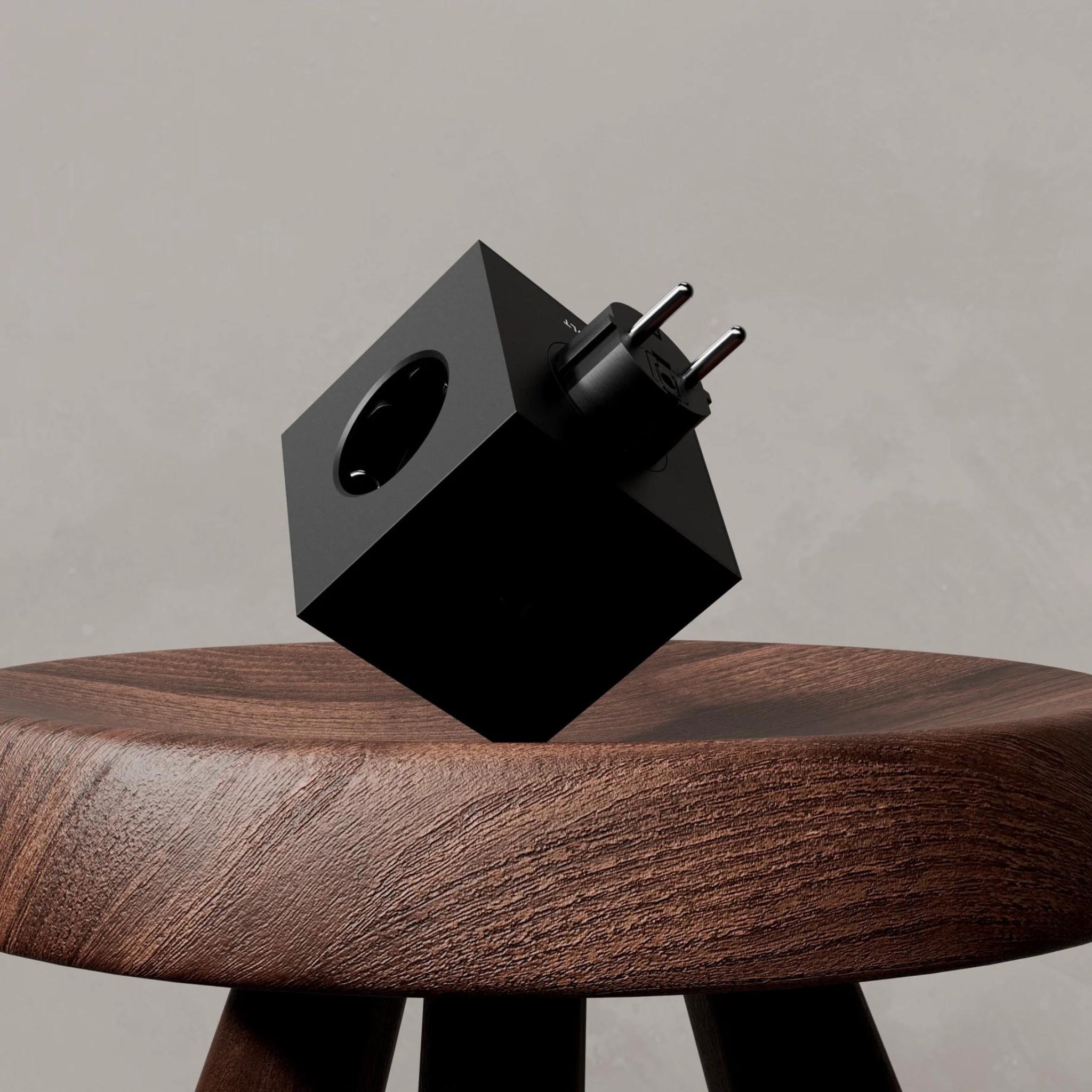 De Avolt Square 2 USB-C Stekkerdoos Stockholm Zwart, een zwarte kubusvormige Schuko-adapter met twee ronde pinnen van Avolt, zit op een houten kruk tegen een neutrale, lichte achtergrond.