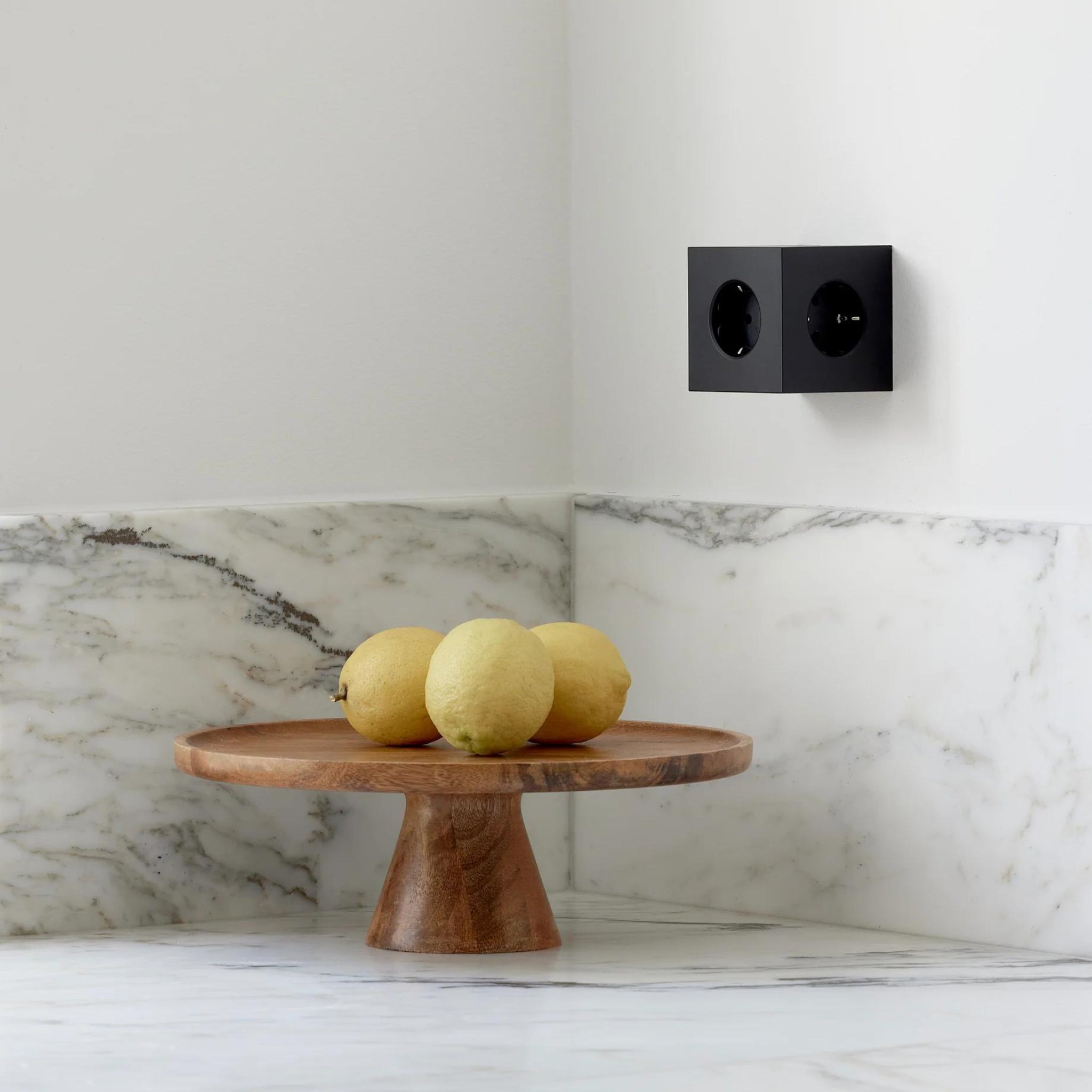 Drie citroenen rusten op een ronde houten sokkel op een marmeren aanrecht, met een wit en grijs marmeren achterwand en een Avolt Square 2 USB-C Stekkerdoos Stockholm Zwart van Avolt erboven.