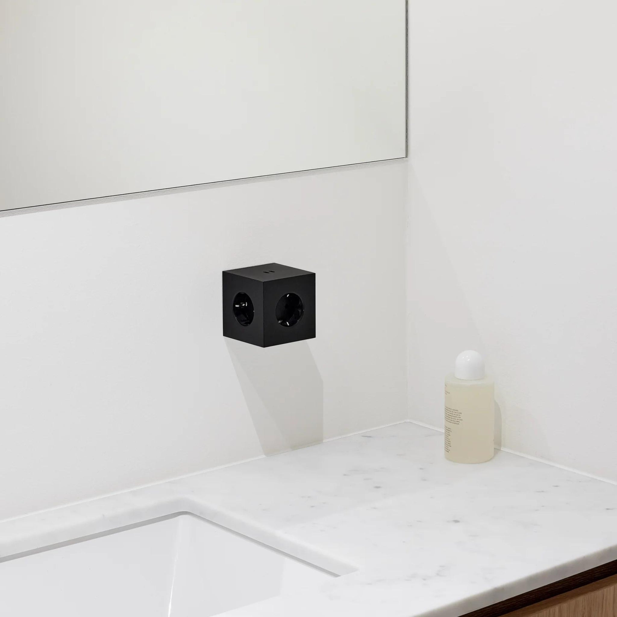 Een minimalistische badkamer met een wit marmeren aanrechtblad, rechthoekige wastafel, fles vloeibare zeep en een Avolt Square 2 USB-C Stekkerdoos Stockholm Zwart stopcontact onder een frameloze spiegel.