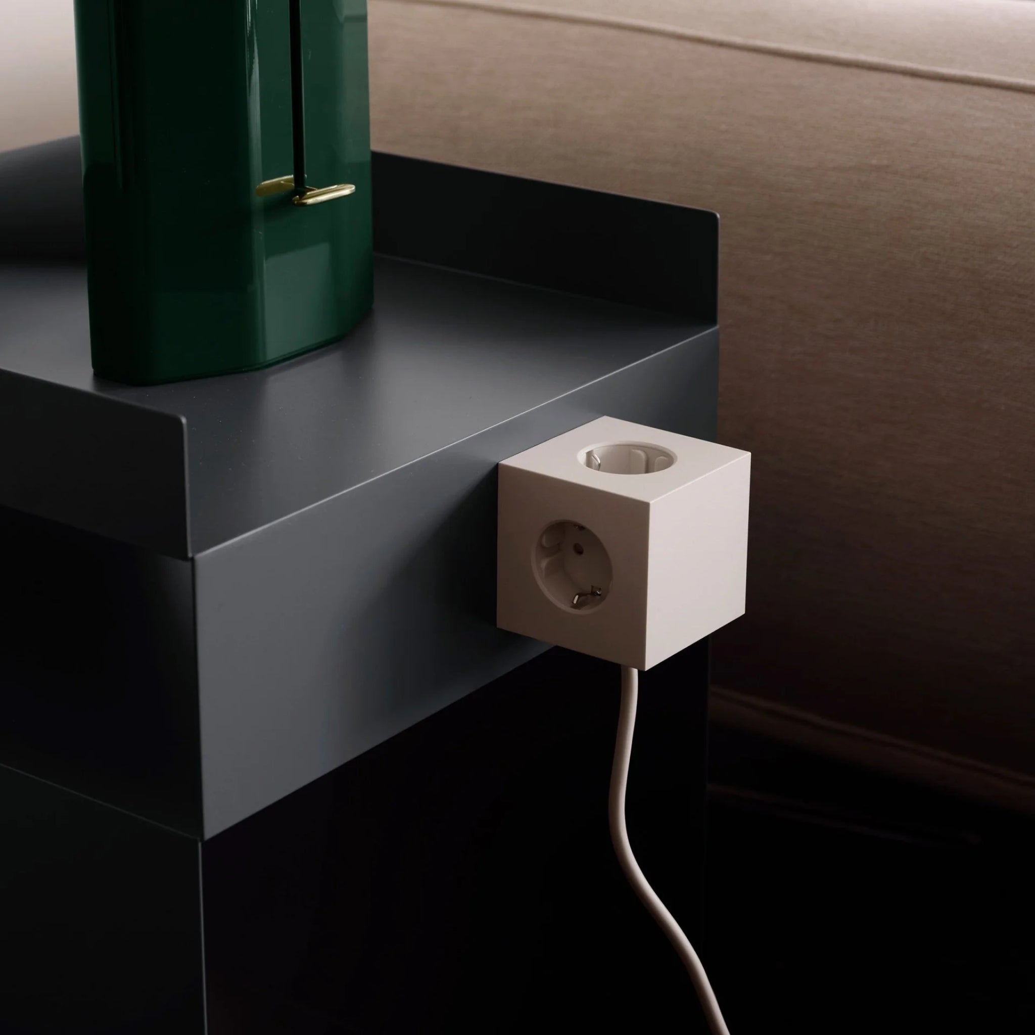 Een Avolt Square 1 USB-C Stekkerdoos Beige 1,8 m is bevestigd aan de zijkant van een donkere tafel bij een beige bank, met een groen object, mogelijk een lamp, bovenop de tafel.