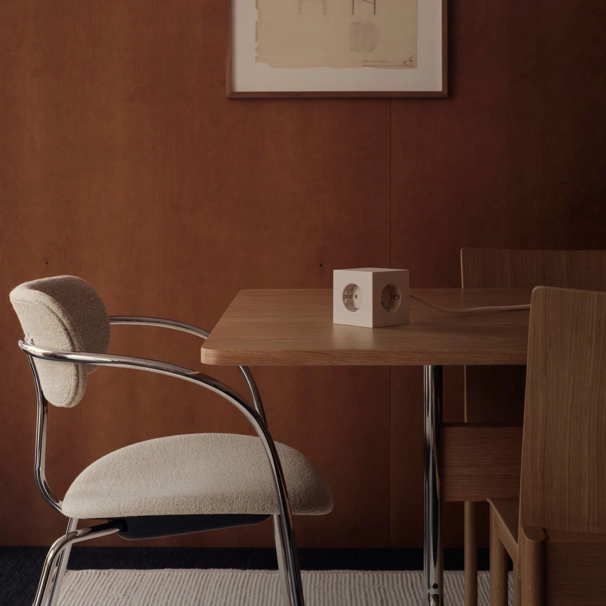 Een moderne eethoek met een houten tafel, minimalistische stoelen en een stoel van chroom en stof is voorzien van een witte kubus Avolt Square 1 USB-C Stekkerdoos Beige 1,8 m om eenvoudig op te laden, abstracte kunst op een bruine muur en een licht vloerkleed op de donkere vloer.