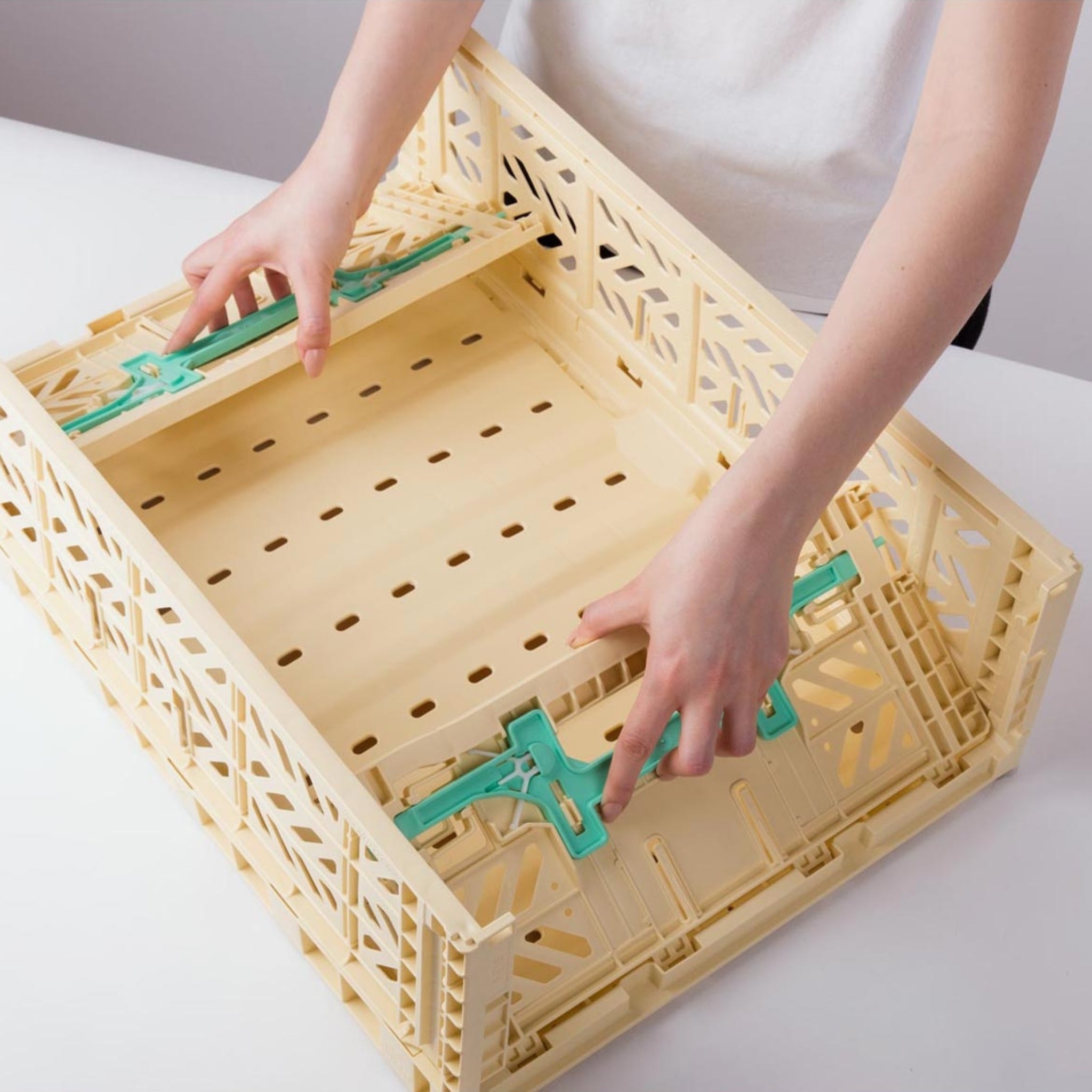 De handen van een persoon vouwen langs de zijkanten van een beige Aykasa Vouwkrat Maxi Comfort Lock Banana / Munt opbergkist van Aykasa, met groene scharnieren, geplaatst op een wit oppervlak.