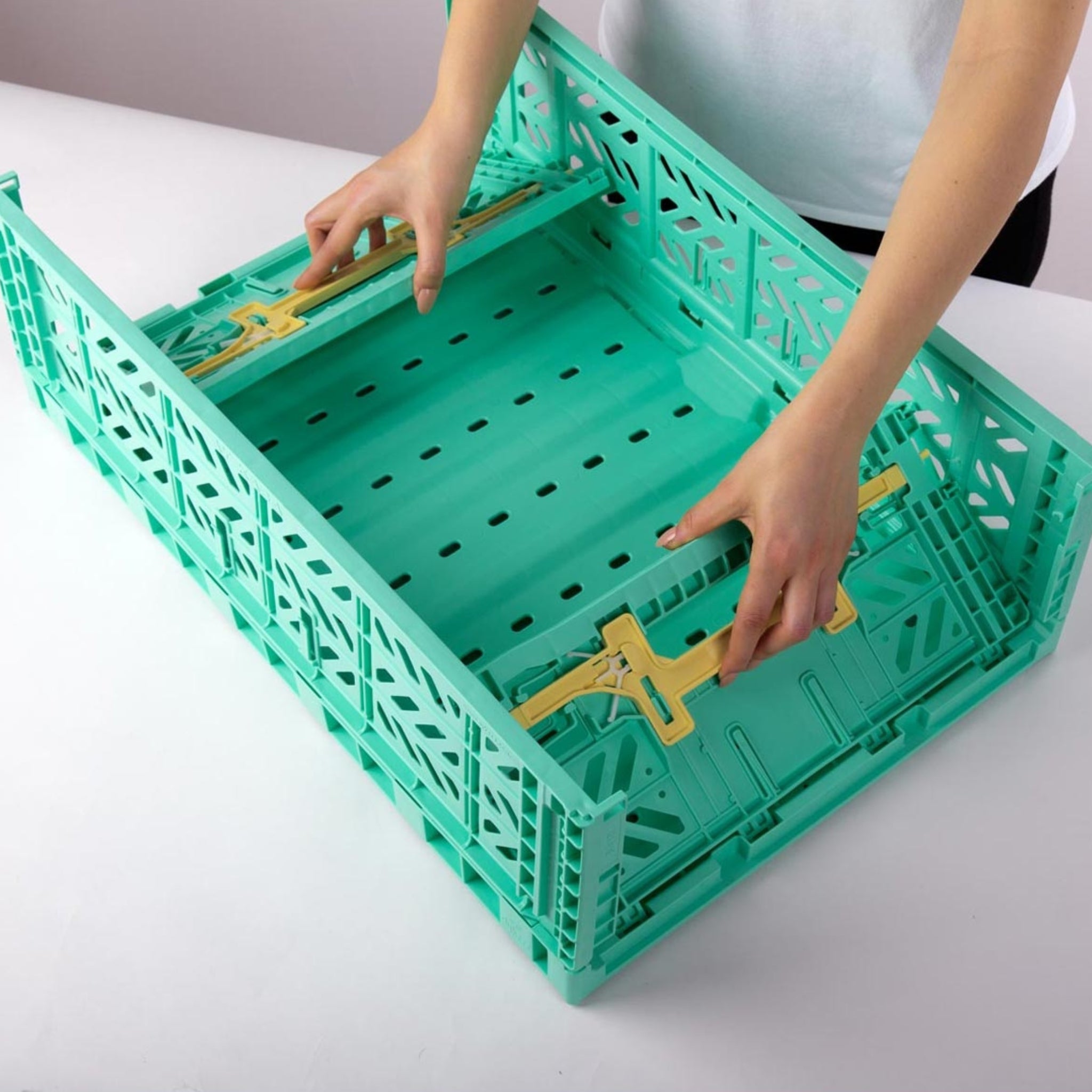 Een persoon zet een Aykasa Vouwkrat Maxi Comfort Lock Munt / Banana opbergkrat van Aykasa in elkaar op een witte tafel.