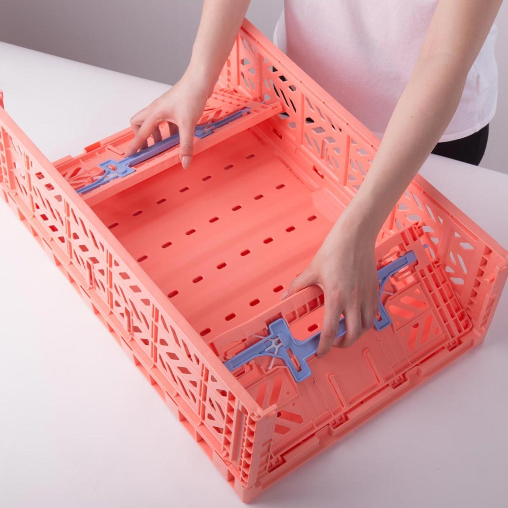 Iemand zet een Aykasa Vouwkrat Maxi Comfort Lock in zalmroze/babyblauw in elkaar door de zijkanten op hun plaats te klikken op een witte tafel.