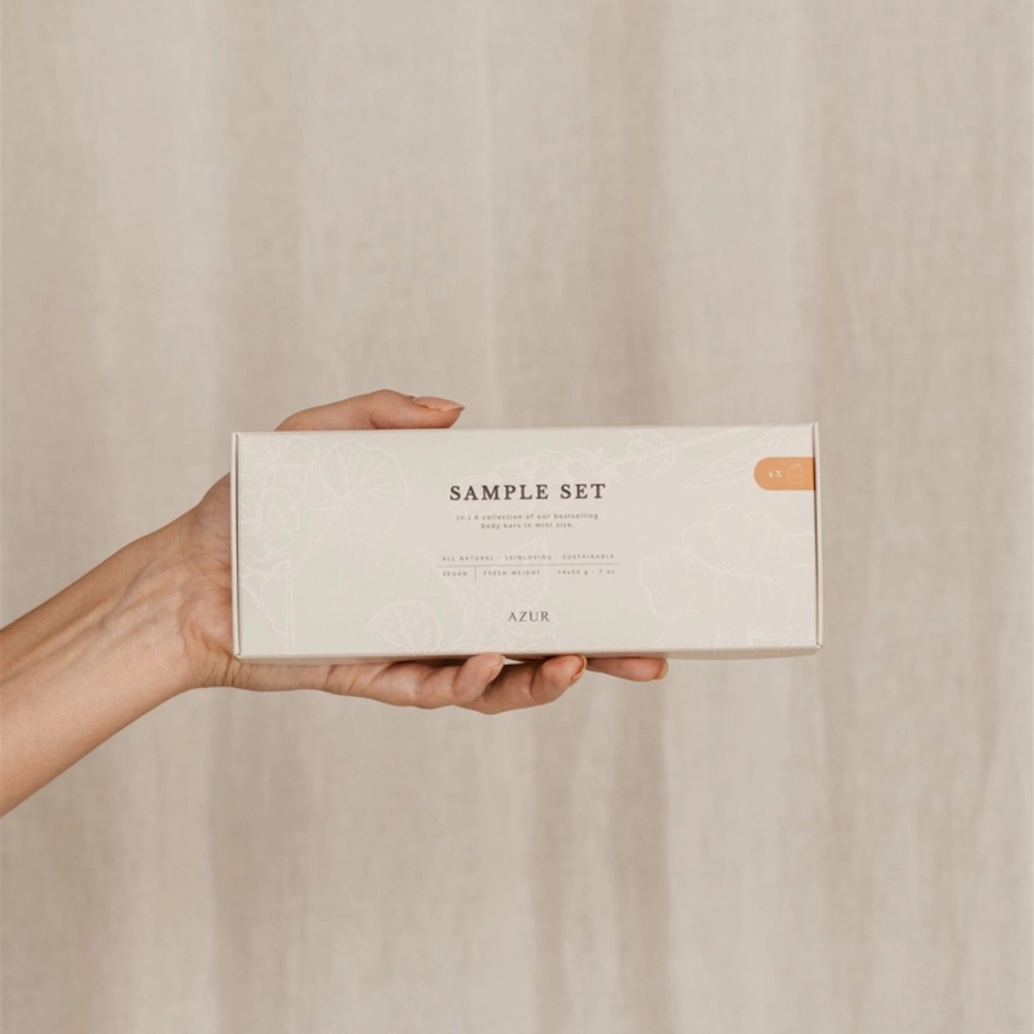 Een hand houdt een rechthoekige beige doos vast met de tekst "Azur Sample Set Natuurlijke zepen 4 st." van Azur, met natuurlijke zepen tegen een neutrale, lichtgekleurde stoffen achtergrond.