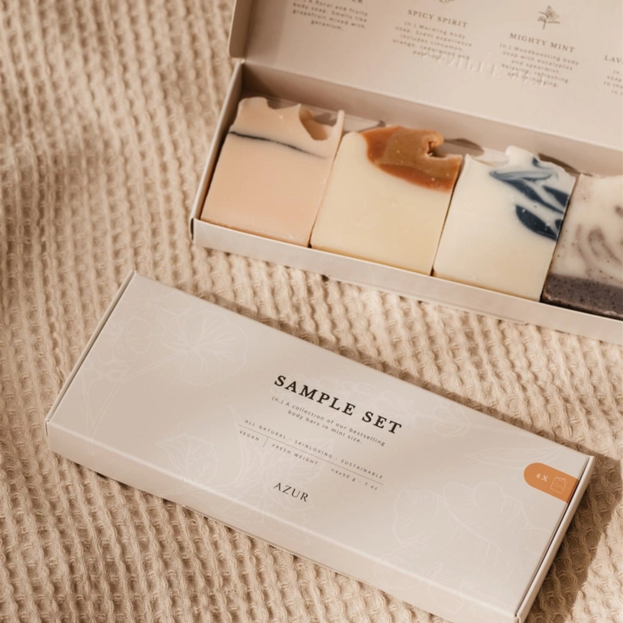 De Azur Sample Set Natuurlijke zepen 4 st. bevat vier plasticvrije, natuurlijke bodybars in verschillende kleuren en patronen. De doos toont de merknaam Azur en is gefotografeerd op beige stof met structuur.