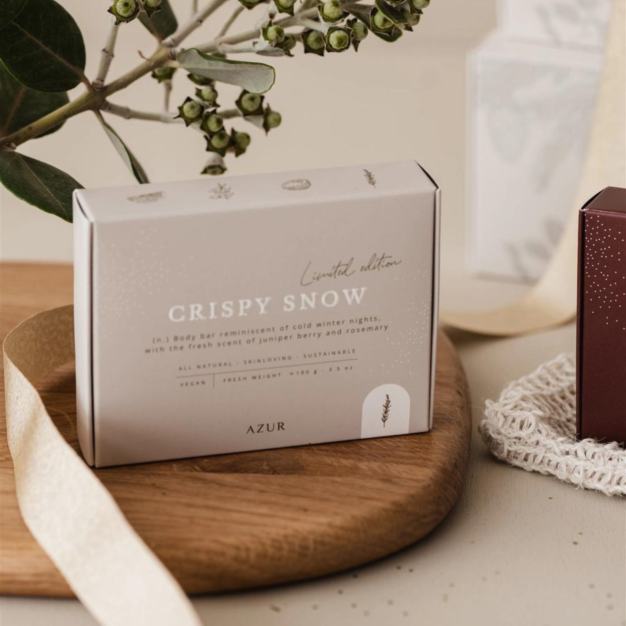 De Azur Crispy Snow - Kerst Natuurlijke Zeep Bar staat op een houten dienblad met gebladerte. De duurzame verpakking benadrukt veganistische, duurzame ingrediënten en een geur van jeneverbes en rozemarijn, bij een kastje in kastanjebruin en een kanten doekje.