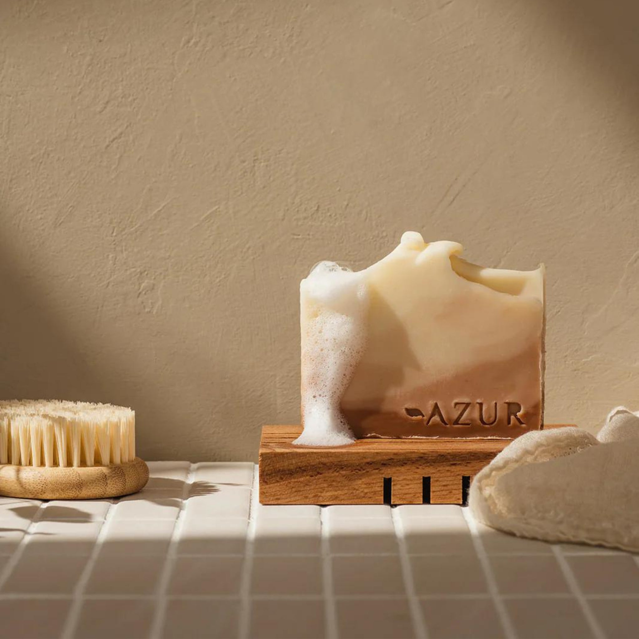 De Azur Houten Zeephouder van Azur, met daarop een zeepbar en schuim, ligt op witte tegels naast een droge borstel en washandje voor een beige muur.