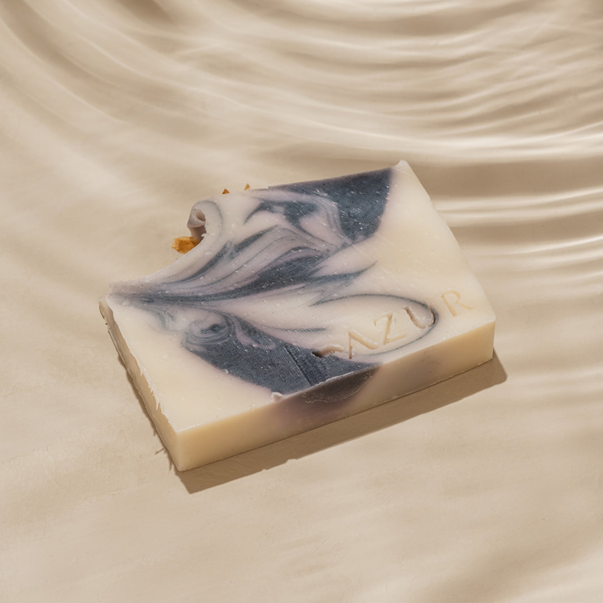 Een rechthoekige Azur Mighty Mint Natuurlijke Zeep Bar van Azur, met witte en donkerblauwe wervelingen en "AZUR" erop in reliëf, rust op een licht golvend oppervlak, met in één hoek een beetmerk.