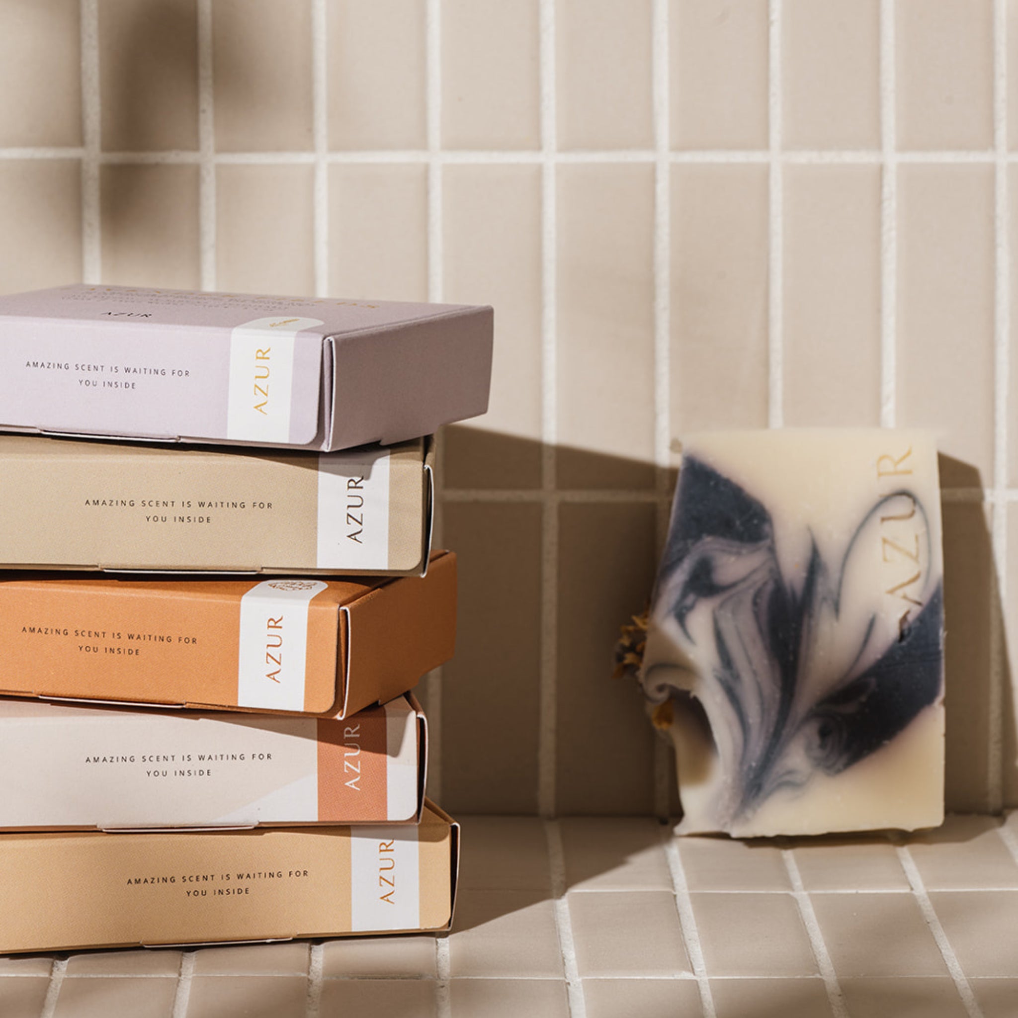 Een stapel van vier pastelkleurige AZUR dozen met duurzame verpakking rust op tegels naast een oningepakte Azur Mighty Mint Natuurlijke Zeep Reep, waarvan de gemarmerde zwarte en witte wervelingen in het zonlicht zachte schaduwen werpen.