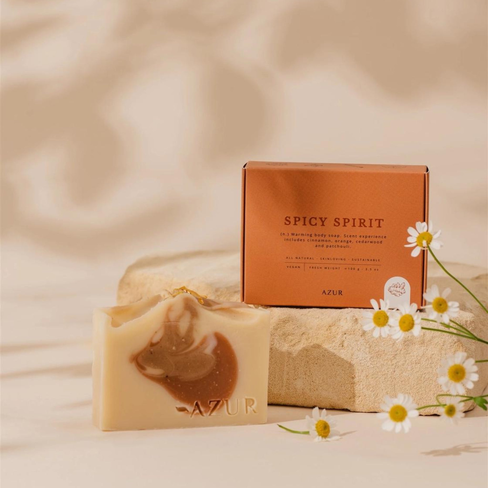 De Azur Spicy Spirit Natuurlijke Zeep Bar rust op beige steen naast zijn oranje doosje. Witte en gele madeliefjes staan op de voorgrond tegen een zachte, neutrale achtergrond. Geïnspireerd door kurkuma.