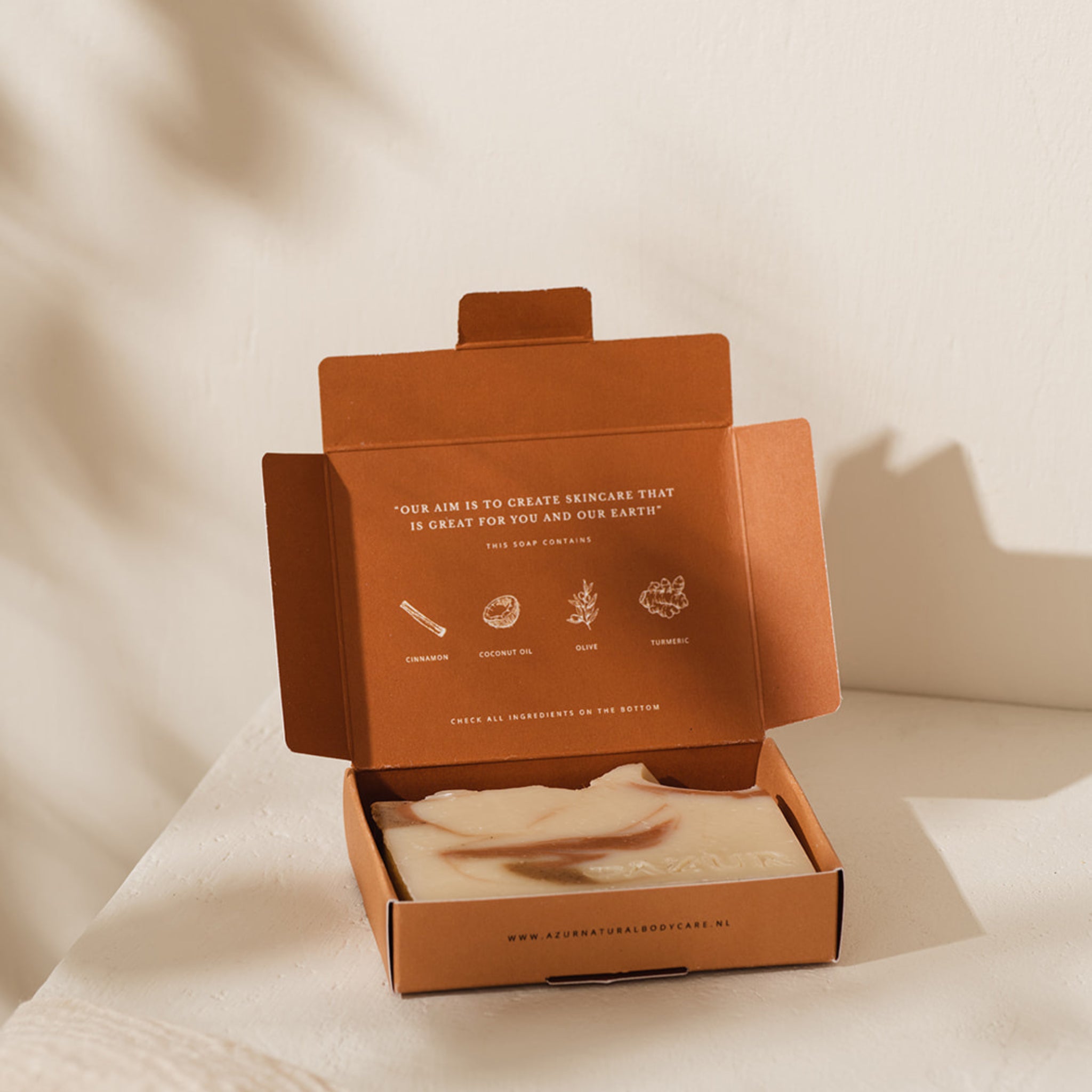 Een open bruine doos met een Azur Spicy Spirit Natuurlijke Zeep Bar zit op een wit oppervlak. De binnenkant van het deksel toont tekst en pictogrammen over de natuurlijke ingrediënten van de zeep van Azur, waaronder voedende kurkuma.