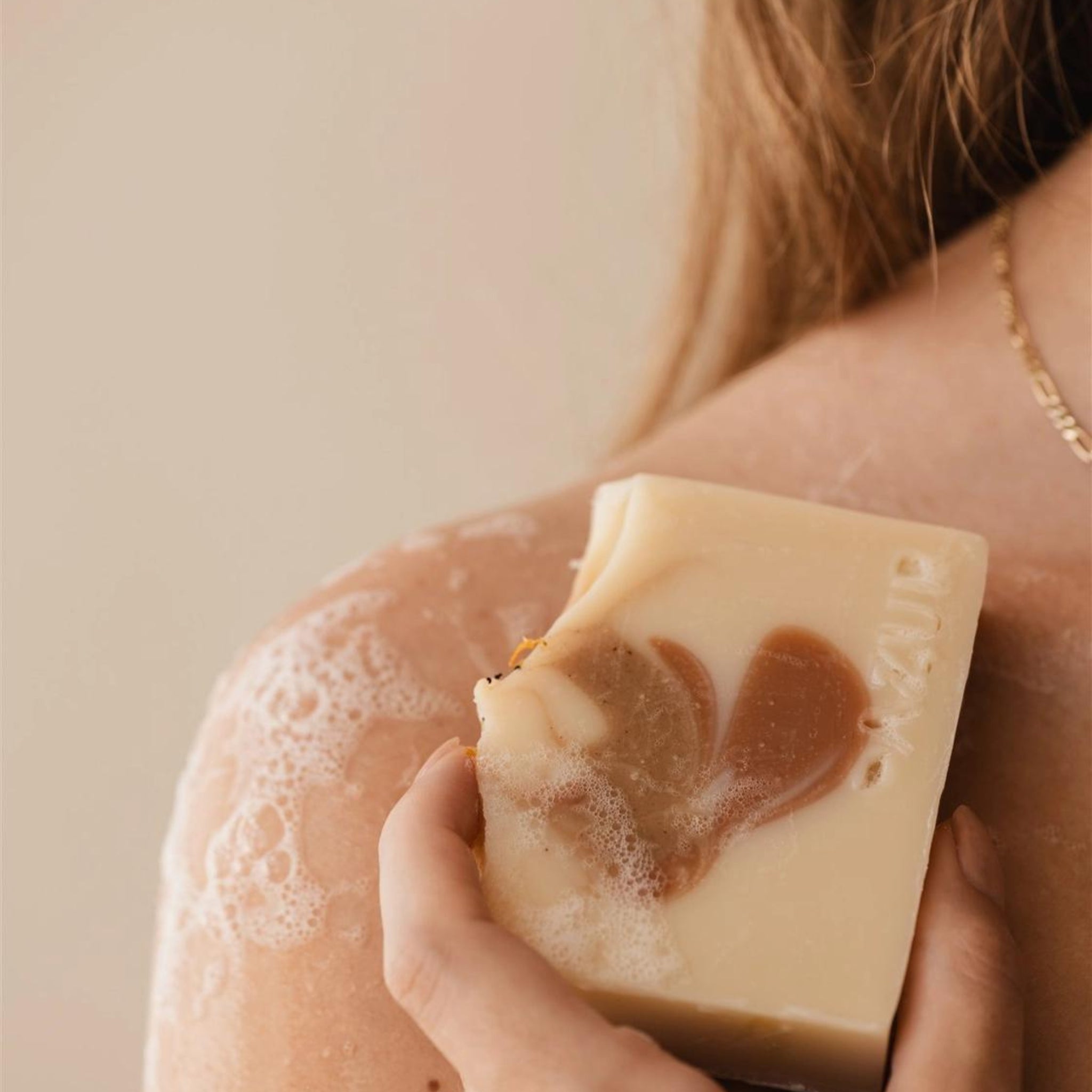 Een persoon met lang haar houdt de Azur Spicy Spirit Natuurlijke Zeep Bar van Azur, met een beige en bruine werveling van kurkuma, tegen zijn zeepschouder. Alleen een deel van de schouder, rug en ketting zijn zichtbaar.