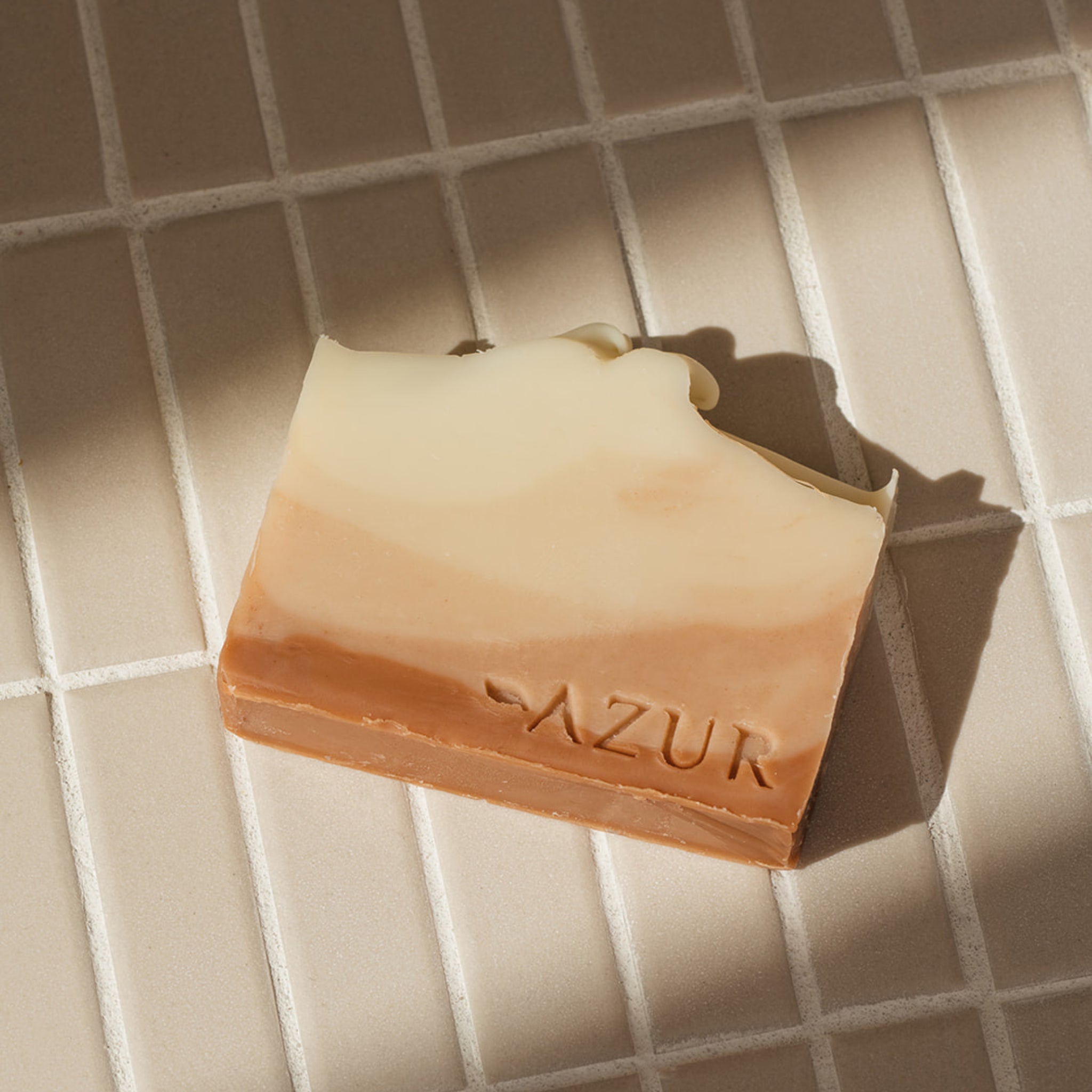 De Azur Sunset Dreams Natuurlijke Zeep Bar, een rechthoekige zeep met crème en bruine lagen met de stempel "AZUR", rust op beige tegels in zonlicht-natuurlijke zeep, handgemaakt in Nederland.