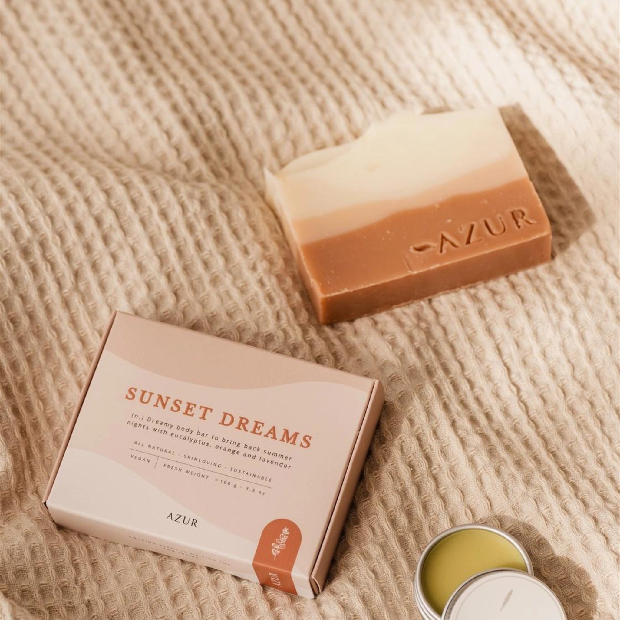 Een Azur Sunset Dreams Natuurlijke Zeep Bar met een roze, beige en bruin kleurverloop rust op beige stof met textuur naast de doos en een open balsemblikje, handgemaakt in Nederland.