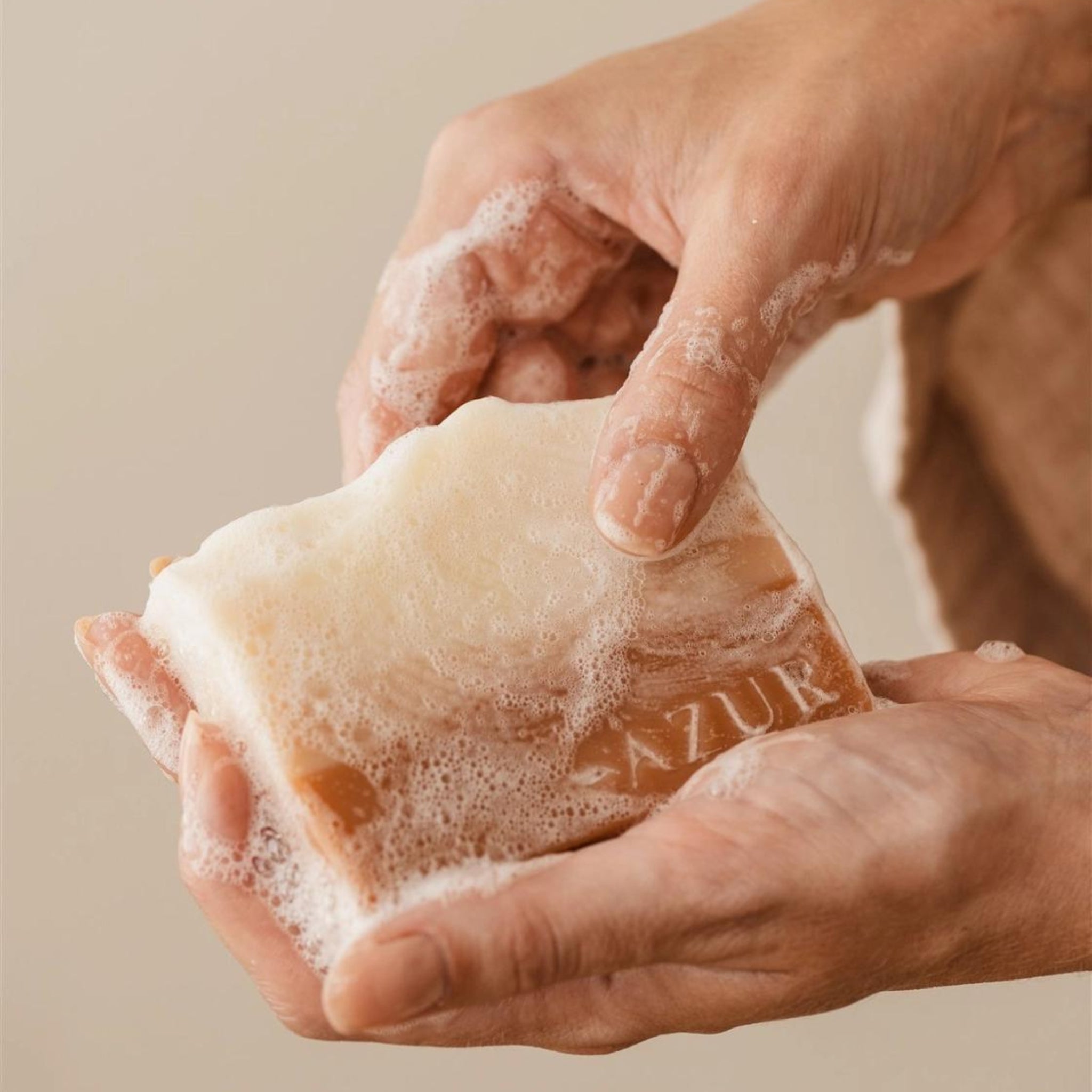 Handen die een Azur Sunset Dreams Natuurlijke Zeep Bar vasthouden, met bubbels en schuim die de bar en vingers bedekken. Het woord AZUR is gedeeltelijk zichtbaar op het oppervlak van de zeep.