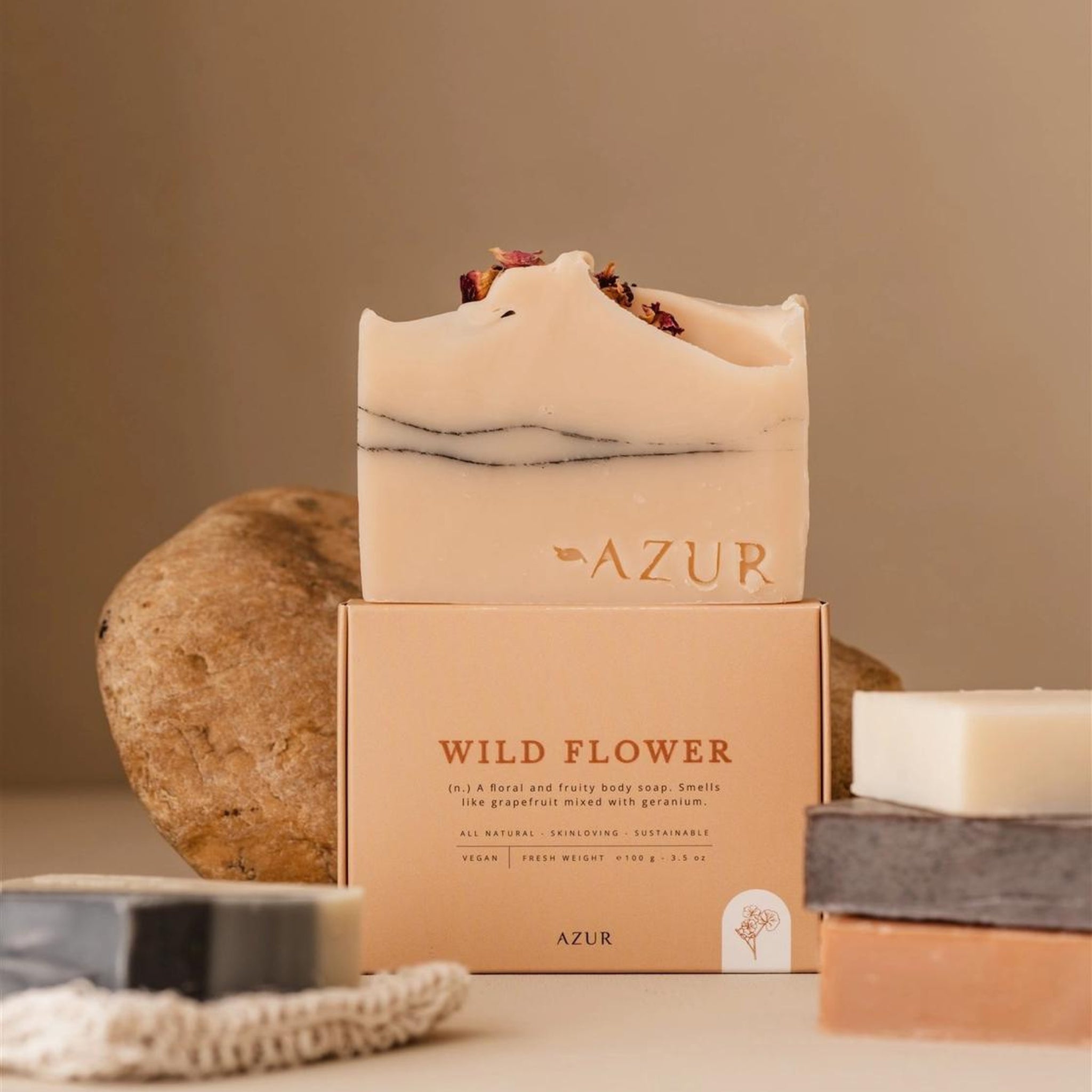 De Azur Wild Flower Natuurlijke Zeep Bar voor droge, gevoelige huid met gedroogde bloemblaadjes ligt op een doosje met Azur-logo, omringd door andere zeep bars en een ruwe steen in een minimalistische, aardse setting.