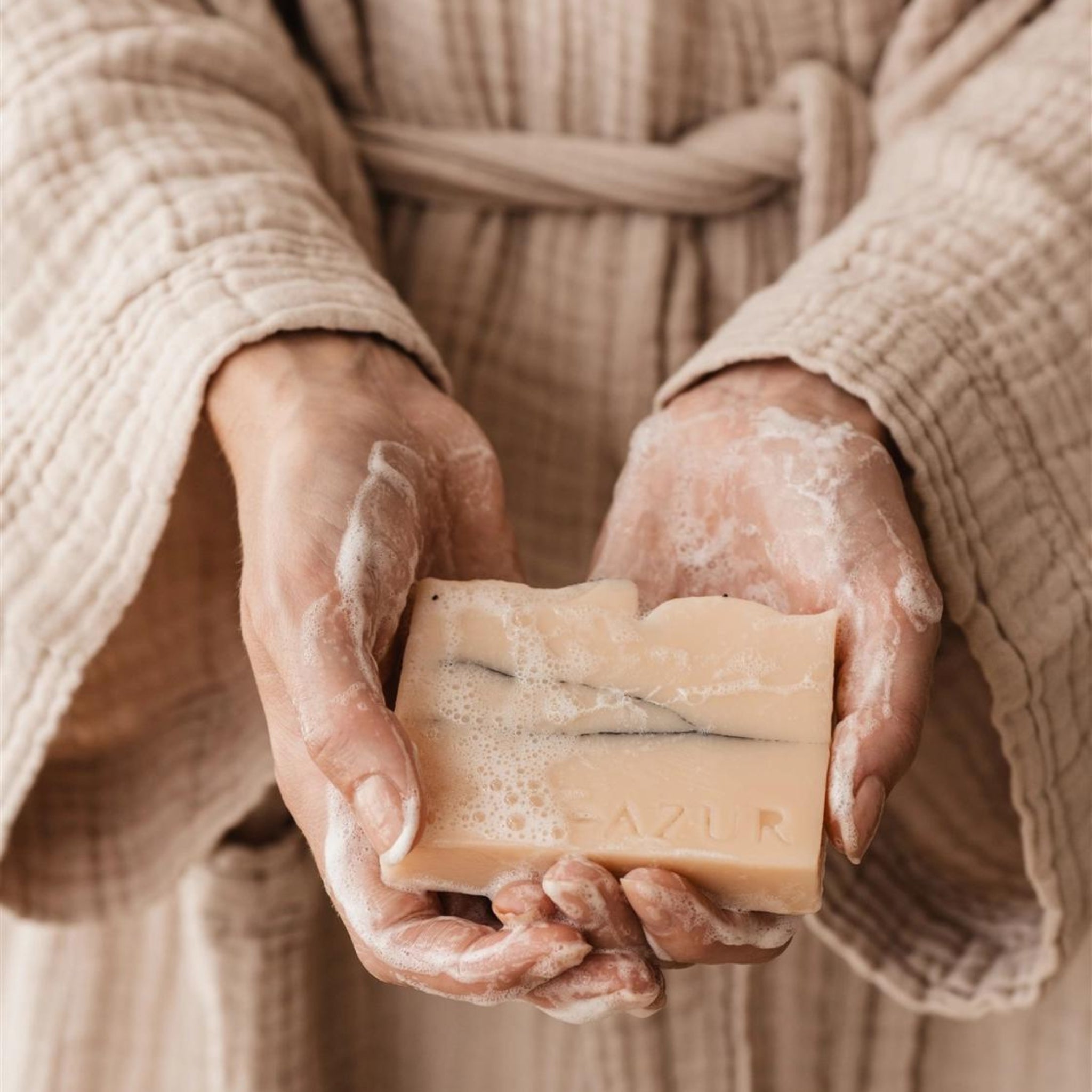 Een persoon in een beige badjas houdt de Azur Wild Flower Natuurlijke Zeep Bar van Azur vast, de naam gegraveerd en natte handen bedekt met zeepsop benadrukken deze zachte zeep die ideaal is voor de droge, gevoelige huid.