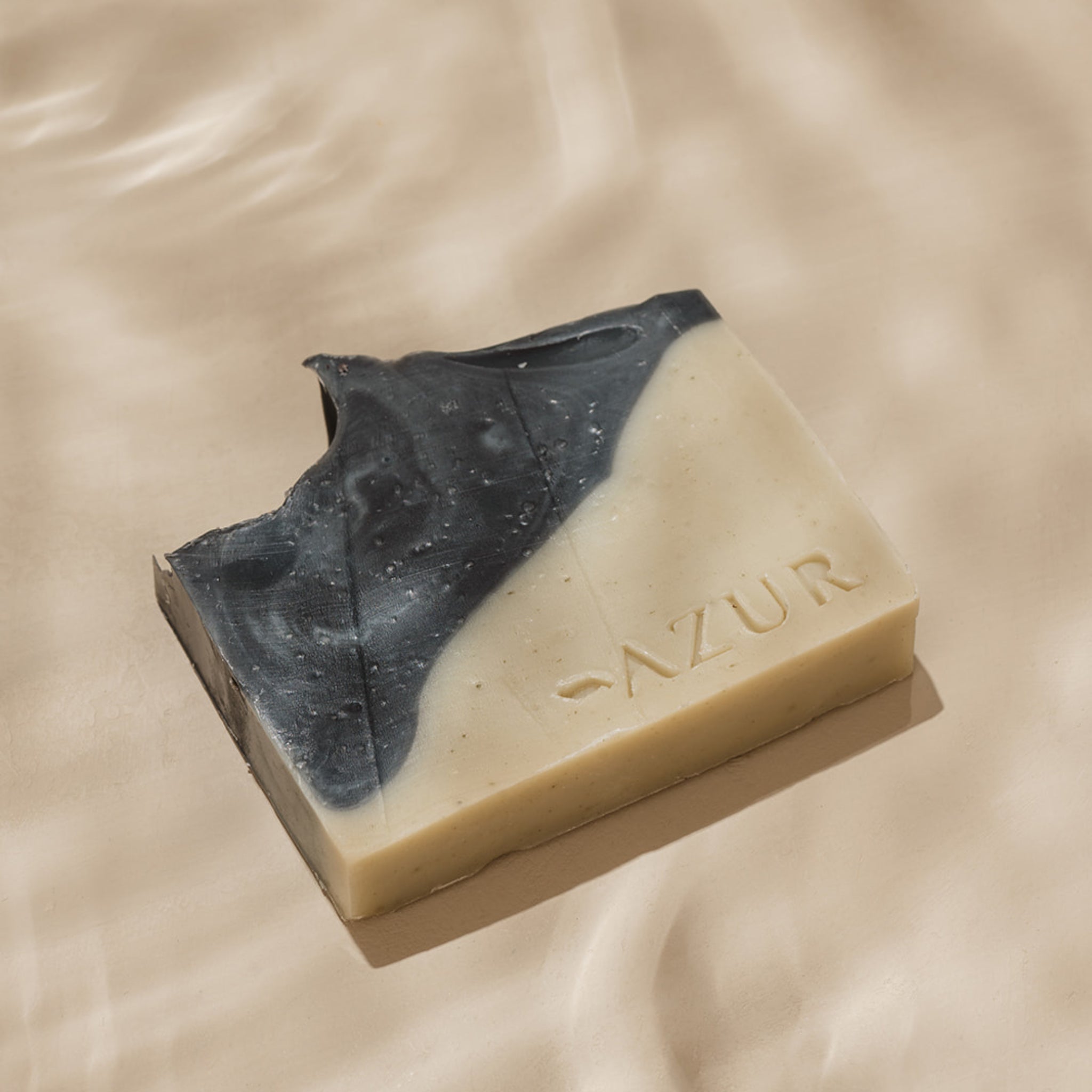 De Azur Wild Woods Natuurlijke Zeep Bar van Azur, met AZUR in reliëf en zwarte en crèmekleurige swirls plus een hapvormige hoek, rust op een glad beige oppervlak en laat ambachtelijke natuurlijke zeep zien.