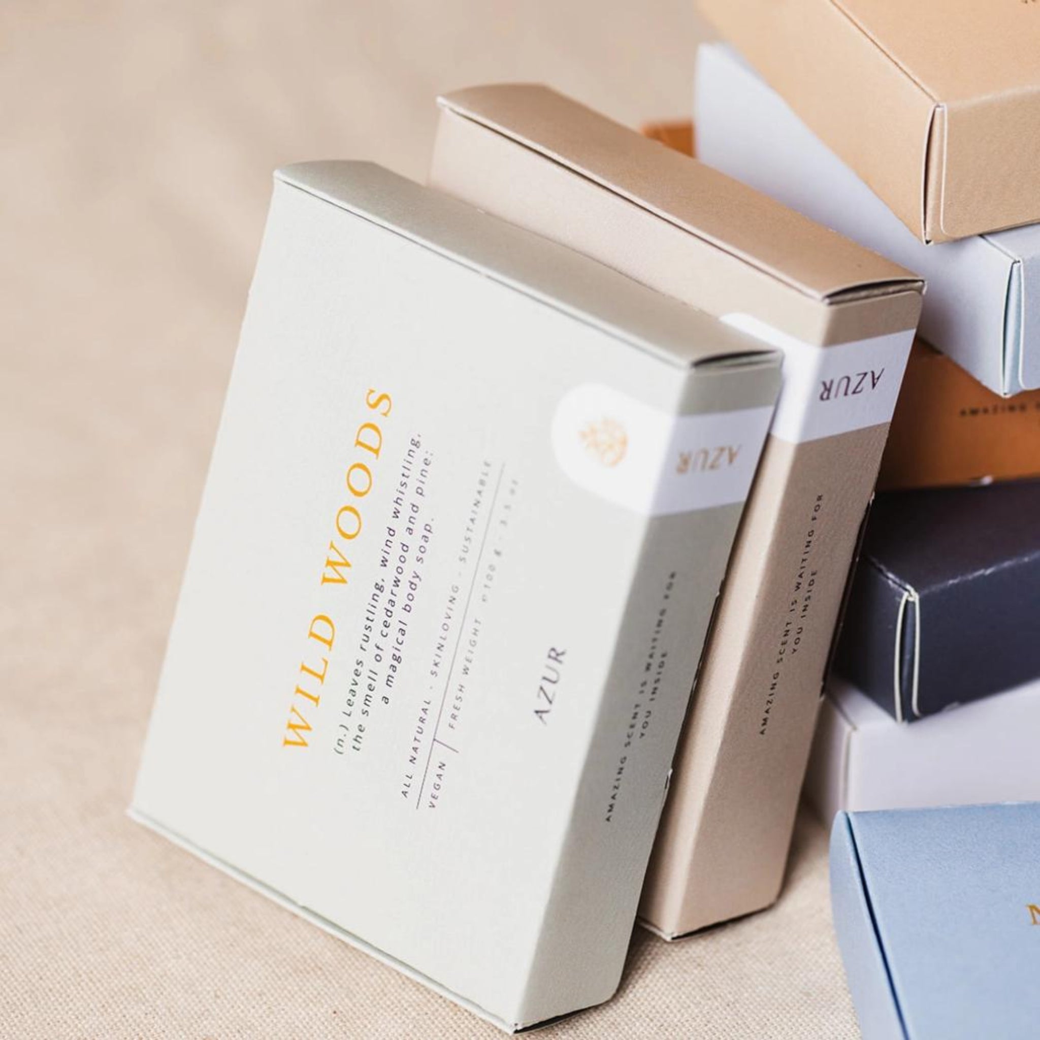 Een close-up van verschillende gestapelde rechthoekige dozen met minimalistische ontwerpen. Op de voorste doos staat de merknaam Azur en de productnaam Azur Wild Woods Natuurlijke Zeep Bar, met de nadruk op duurzame verpakkingen op een beige oppervlak in zachte, neutrale kleuren.