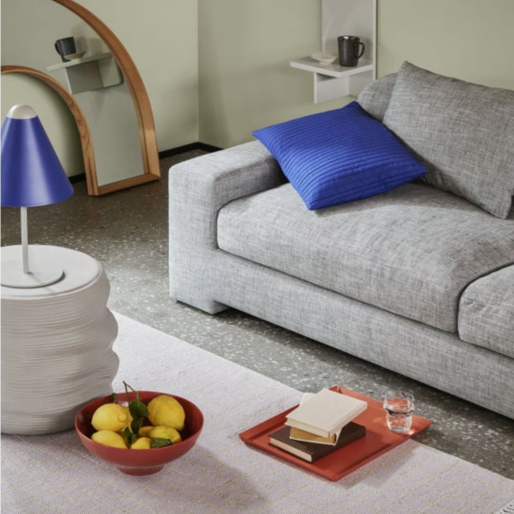 Een moderne woonkamer met een lichtgrijze bank, een blauw kussen, een wit tafeltje met een blauwe lamp, een schaaltje citroenen, boeken en een glas water op het Broste Copenhagen Erika Dienblad Bruin dienblad, gestyled met chique decoratie-objecten.