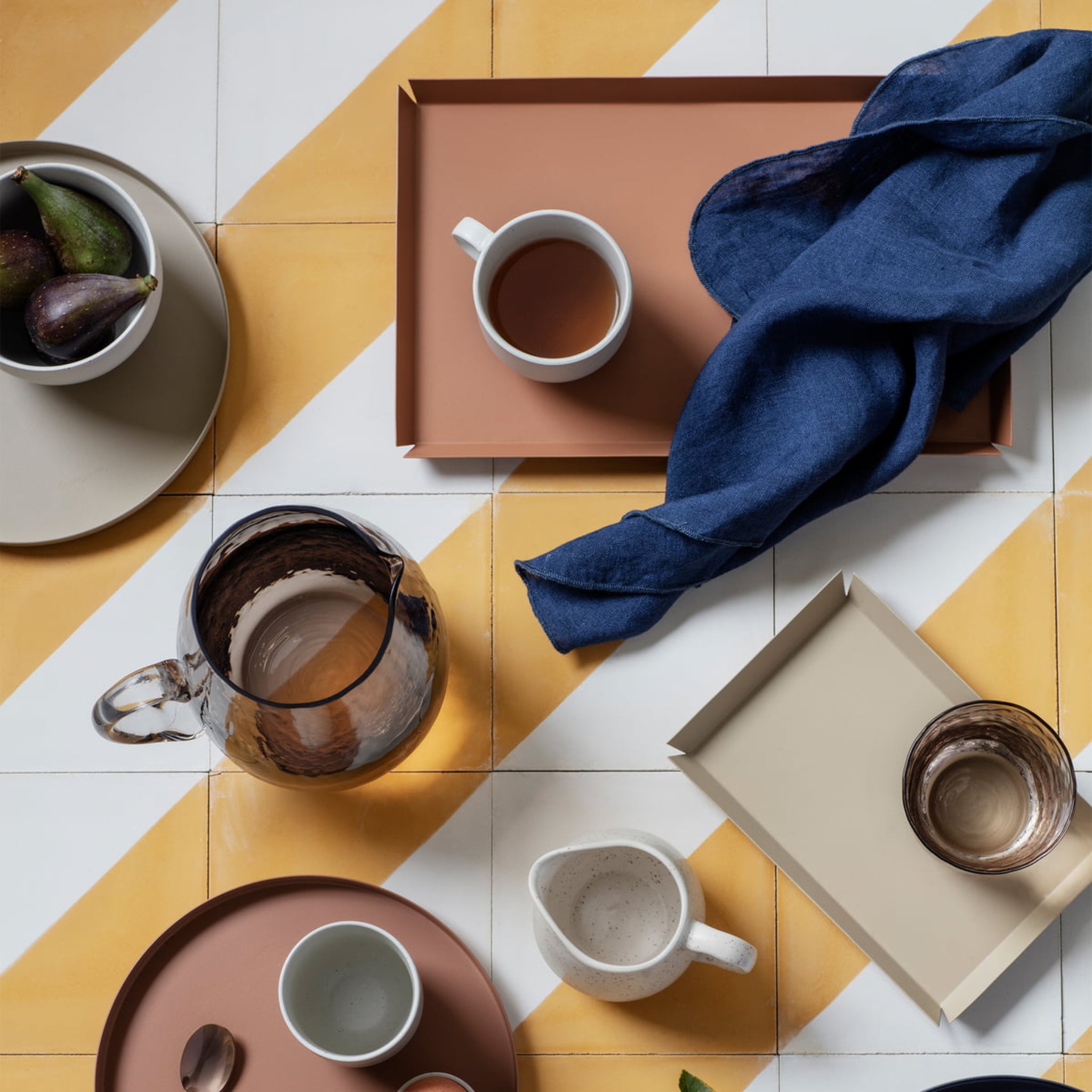 Een bovenaanzicht van een gedekte tafel met dienbladen, mokken, de Broste Copenhagen Erika Dienblad Bruin 33/21, een schaal vijgen en een marine servet op geel en wit gestreepte tegels in een moderne, minimale stijl.