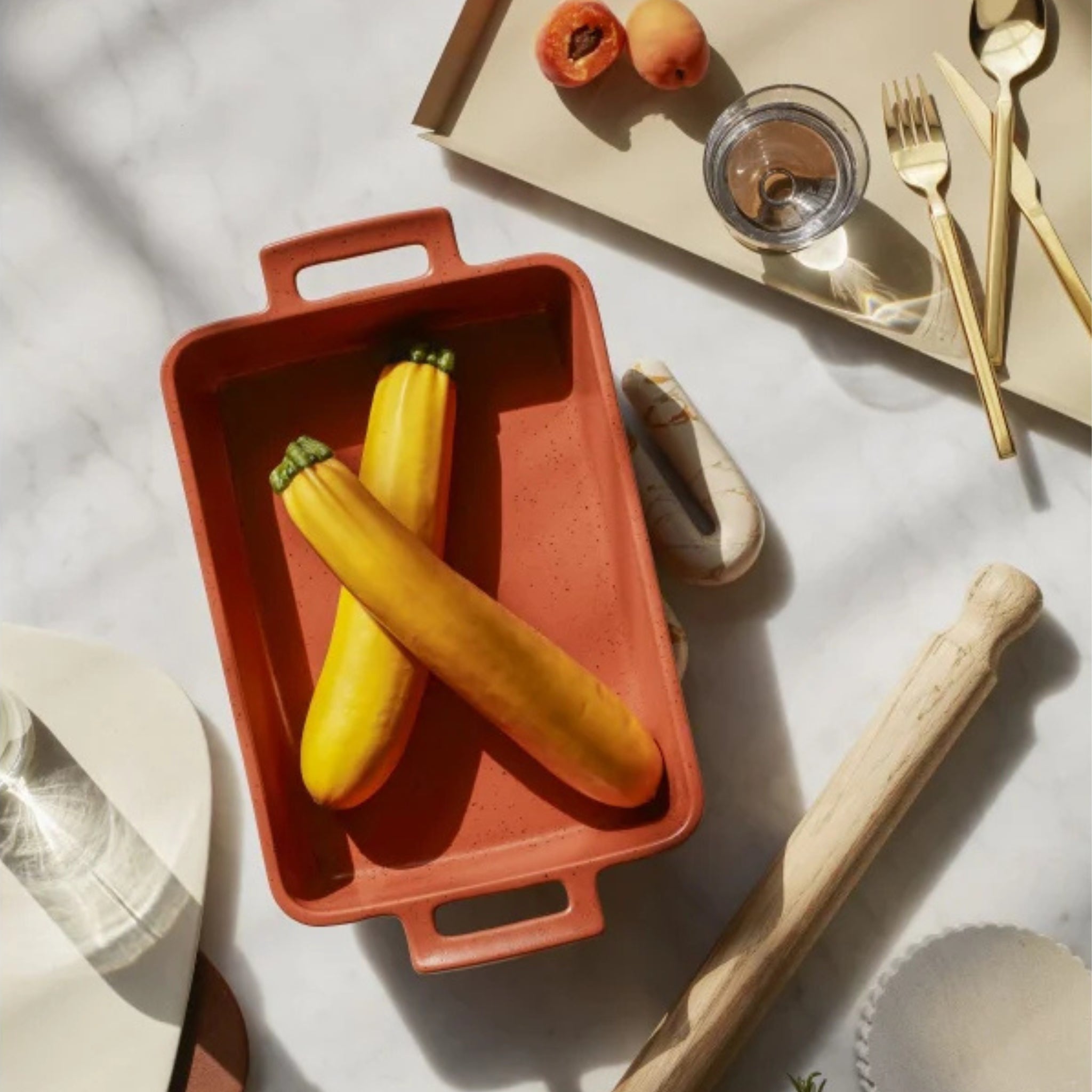 Een Broste Copenhagen terracotta ovenschaal met twee gele courgettes in een X staat op marmer. Vlakbij: een Broste Copenhagen Erika Dienblad Zand 43/33, deegroller, bord, glas, bestek en abrikozen in zacht natuurlijk licht.