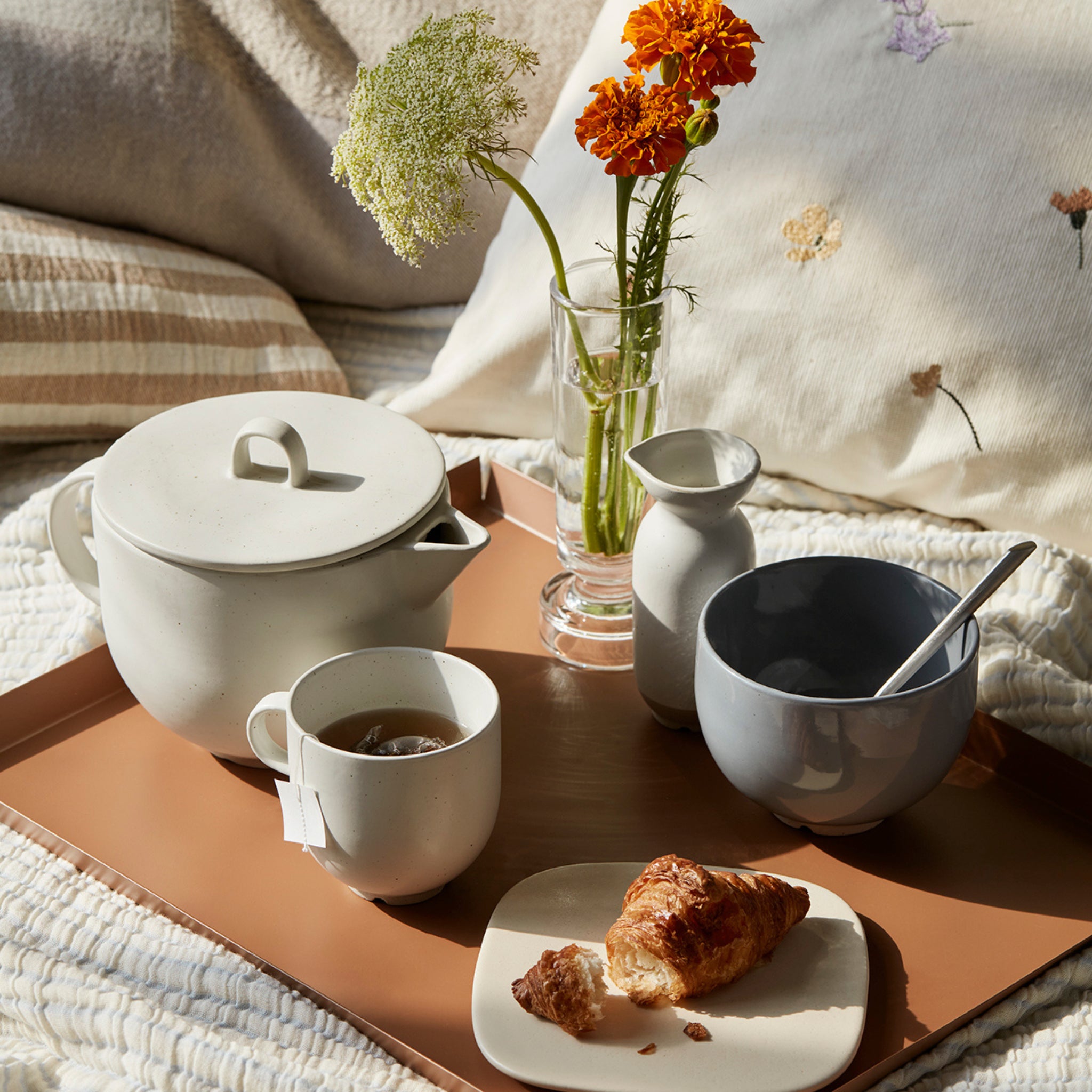 Het Broste Copenhagen Erika Dienblad Zand 43/33 ontbijtblad rust op een bed, met daarop een theepot, theekopje, croissantbord en oranje bloemen in een vaas. Het uitnodigende tafereel wordt gecompleteerd door pluchen kussens en knusse dekens.