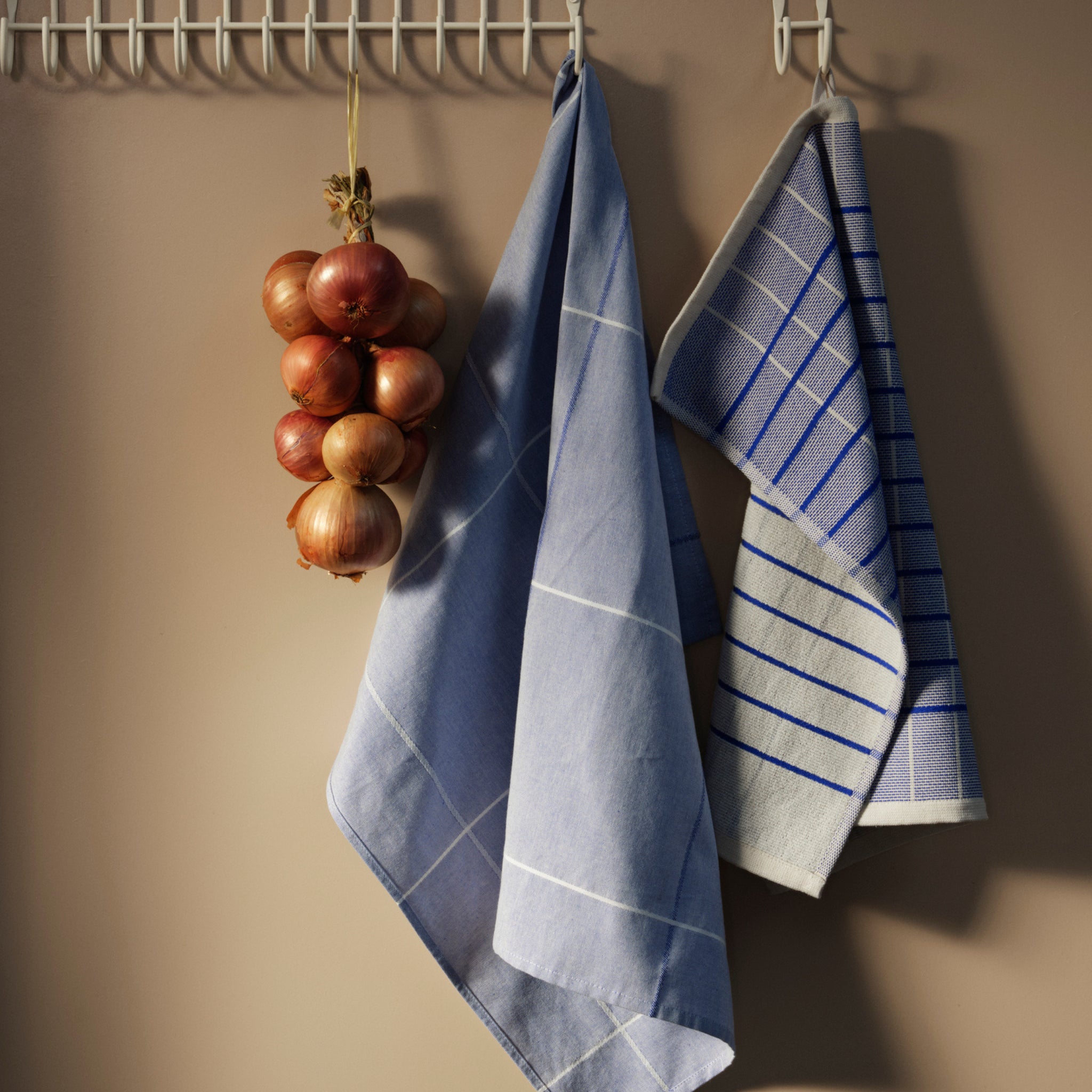Een bos uien hangt aan een wit rek naast twee Broste Copenhagen Herman Theedoeken Blauw (set van 2) tegen een beige muur.