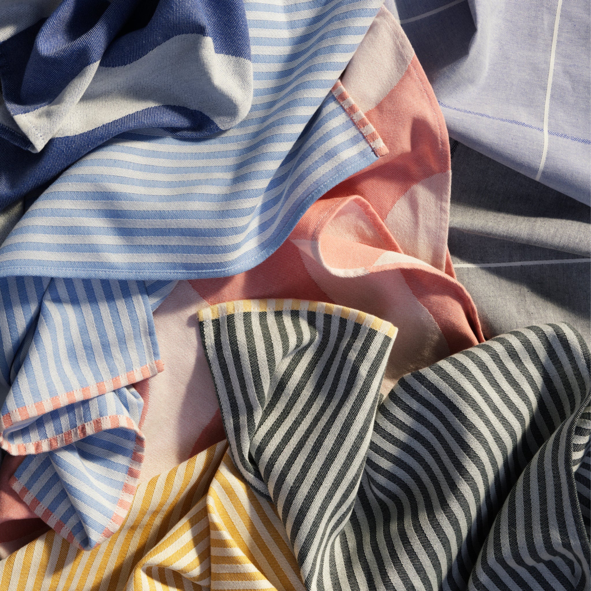 Verschillende blauwe, roze, grijze en gele stoffen - vergelijkbaar met Herman Theedoek Blauw 2 st. van Broste Copenhagen - laten gelaagde gestreepte patronen zien die zacht worden verlicht door natuurlijk licht.