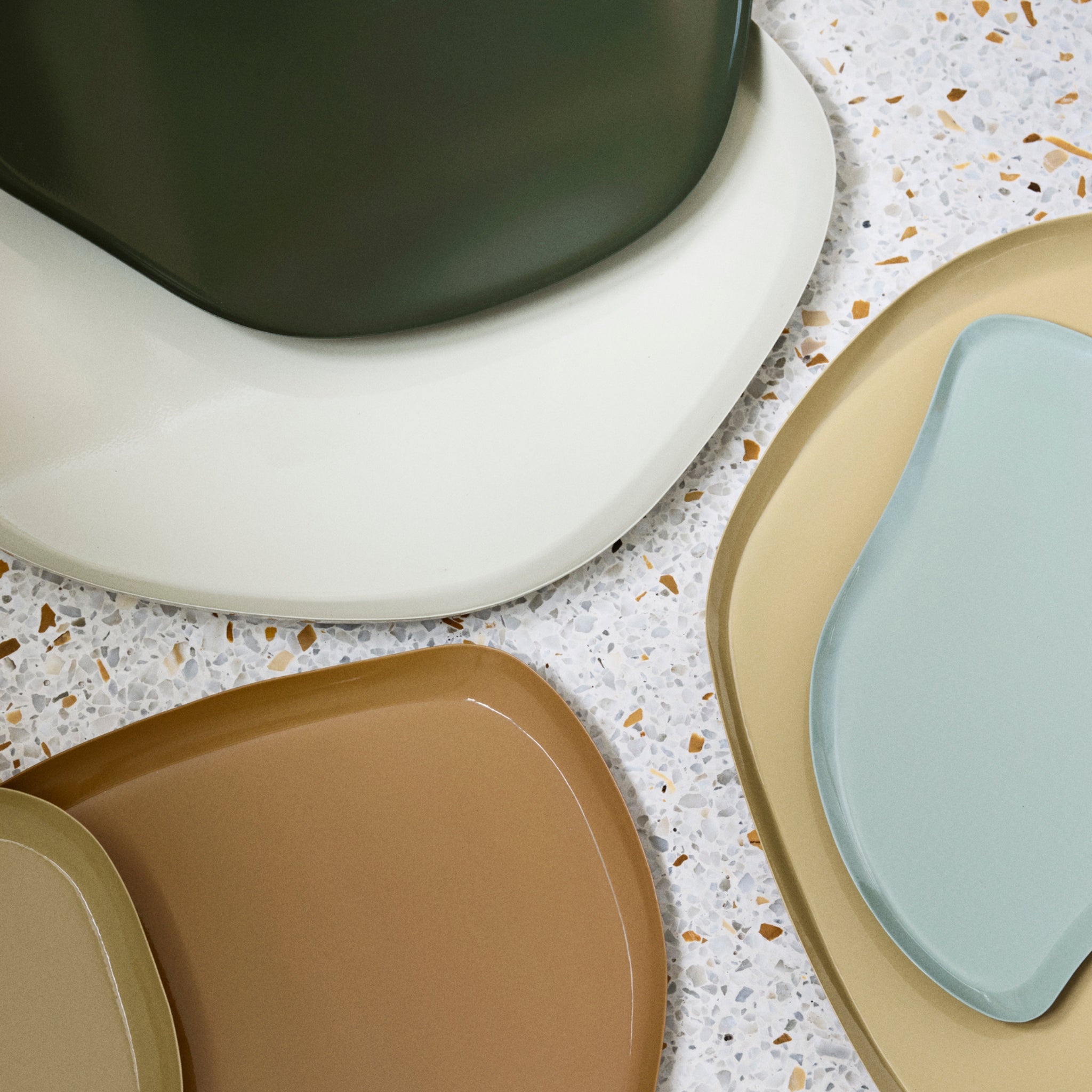 Een close-up toont overlappende, onregelmatig gevormde borden in groen, crème, tan en blauw op een gespikkeld oppervlak, wat het unieke ontwerp van de Broste Copenhagen Mie Dienblad L Ecru 39/32 benadrukt.