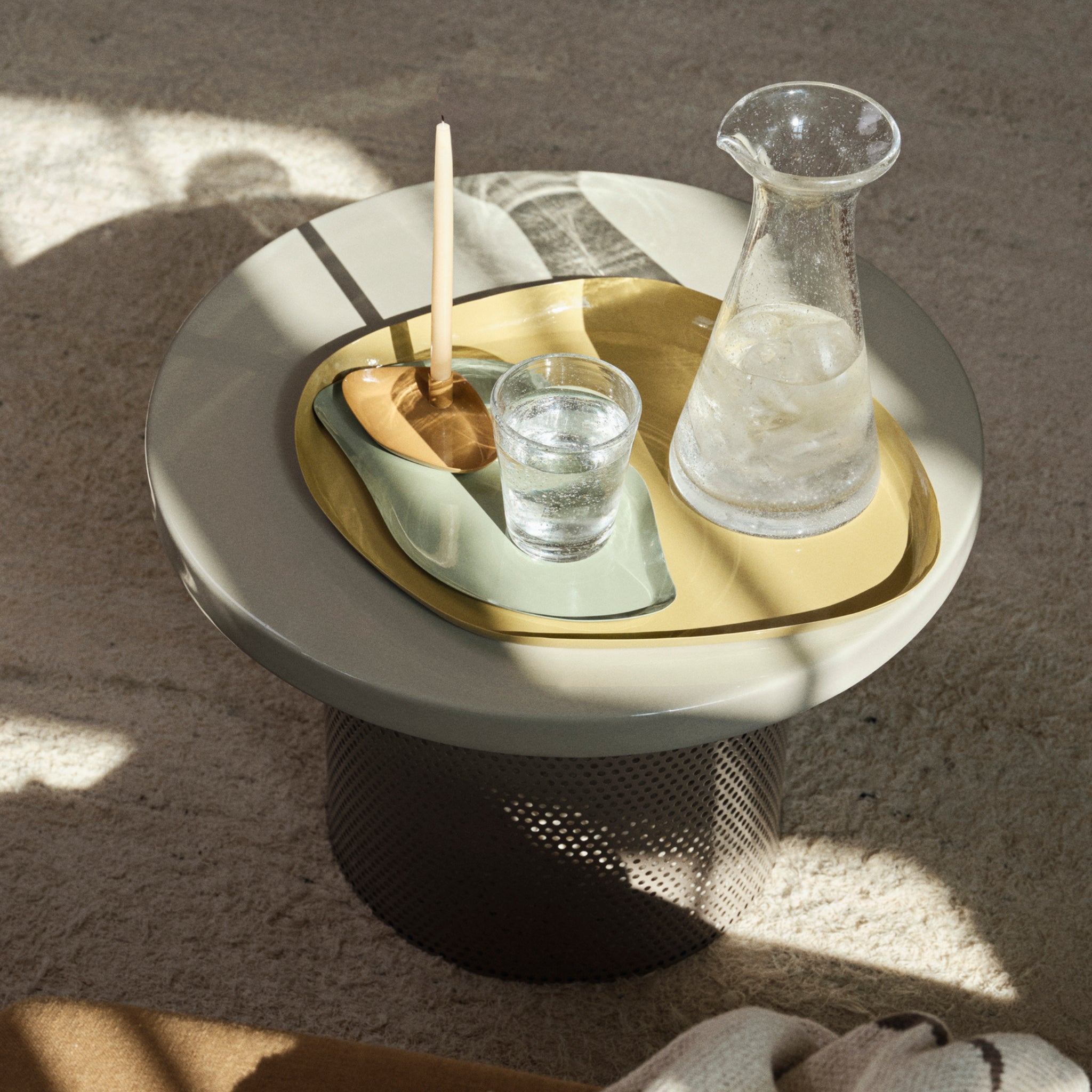 Een rond, lichtgrijs bijzettafeltje toont een geel dienblad met een glas water, een karaf met helder ijswater, een houten lepel en het Broste Copenhagen Mie Dienblad L Taupe Zand 39/32. Zonlicht en schaduwen voegen warmte toe aan het uitnodigende tafereel.