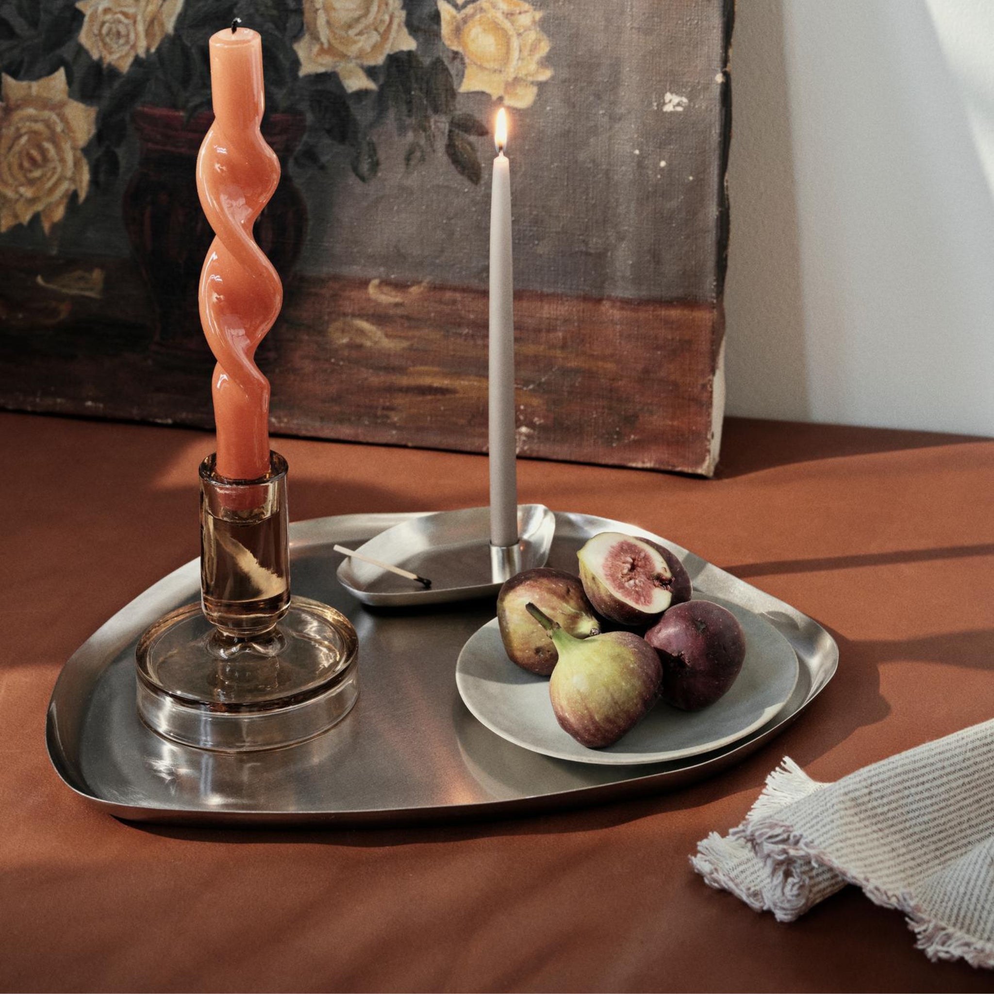 De Broste Copenhagen Mie Dienblad M Zilver 34/27 heeft een oranje gedraaide kaars in een glazen houder en een slanke witte kaars, naast een bord met gesneden vijgen en een doek, alles tegen een bloemenschilderij.