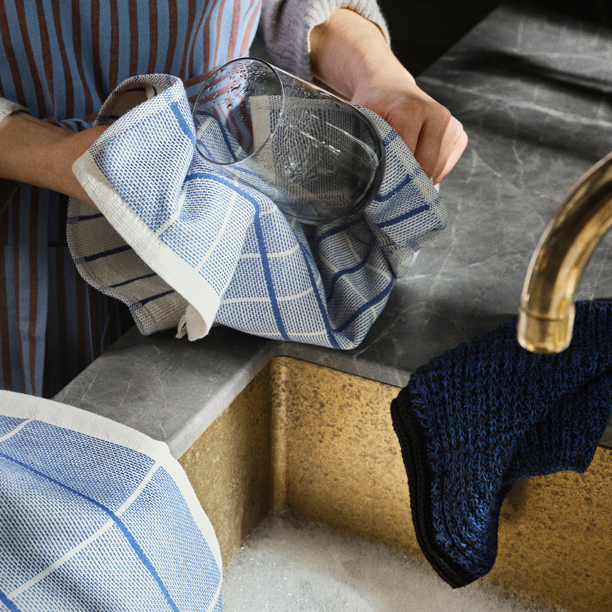 Een persoon droogt een helder glas met een Broste Copenhagen Rigmor Vaatdoeken Blauw boven een gootsteen met zeepsop, met een gestreept schort. Een donkerblauw katoenen washandje hangt op het aanrecht.