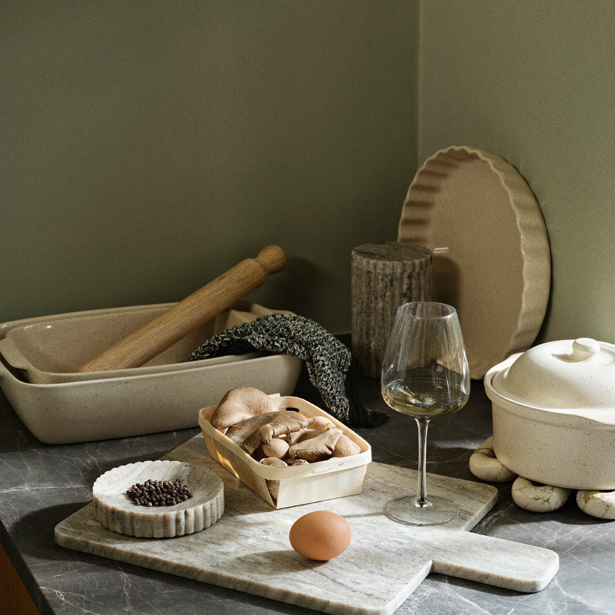 Een keukenscène met een Broste Copenhagen Broste Vig Ovenschaal Beige 25,5/17, een deegroller, een theedoek, een mandje champignons, een wijnglas, een peperbakje, een bruin ei en een pot met deksel op een marmeren aanrecht.