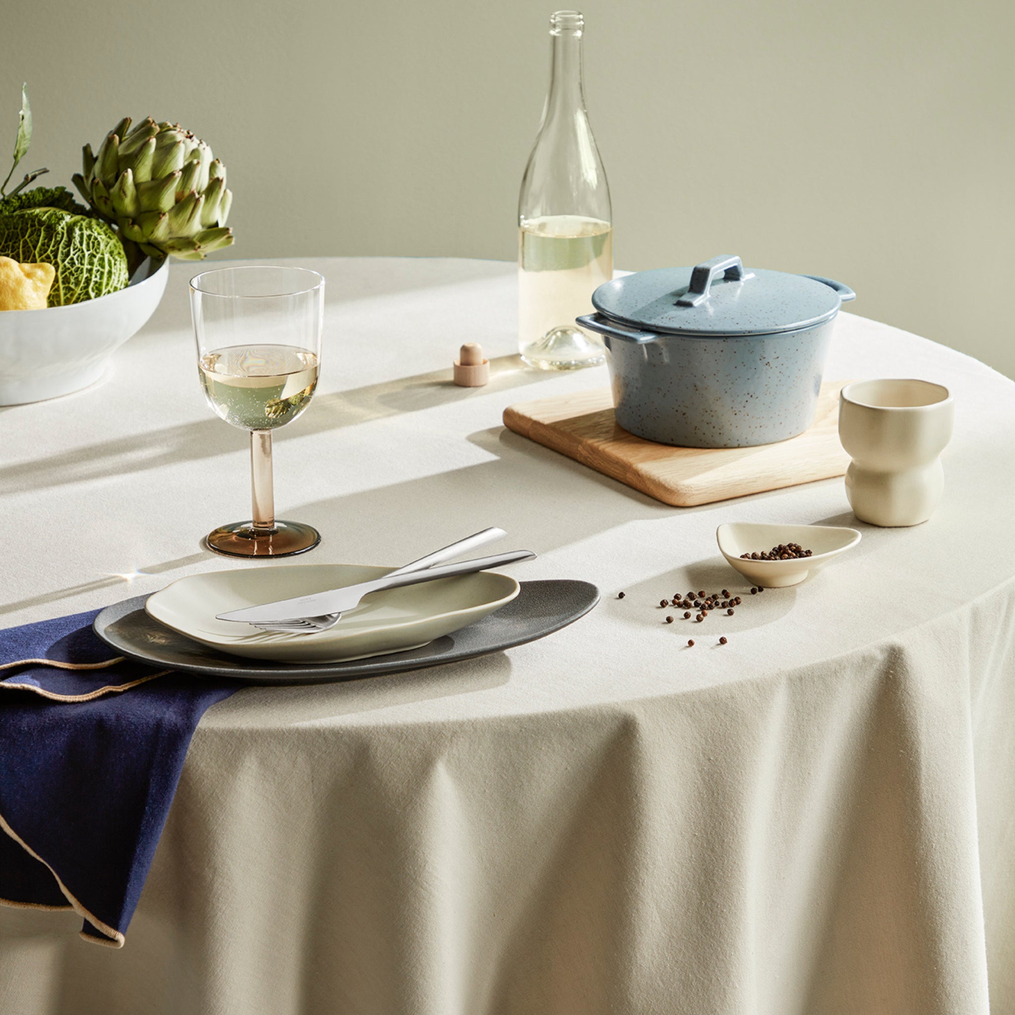 Een ronde tafel met een wit tafelkleed, gedekt voor één persoon, met een bord, vork, mes, Broste Copenhagen Wilhelmina stoffen servet donkerblauw D45, wijnglas, kopje, kommetje met peperkorrels, blauwe pot op een houten plank, fles wijn en kom met groenten.