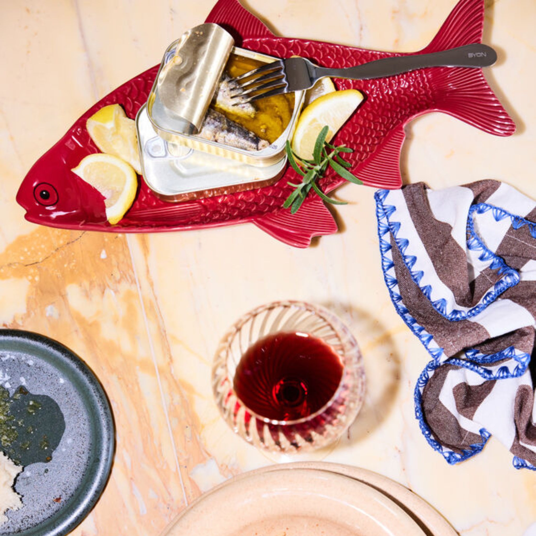 Een Byon Kai Serveerschaal Rood van Byon in de vorm van een vis toont een open blikje sardines met citroenpartjes en kruiden. Een vork rust op het blikje. Vlakbij: een glas rode wijn, zwart bord met kruimels, servet en beige bord op marmer.