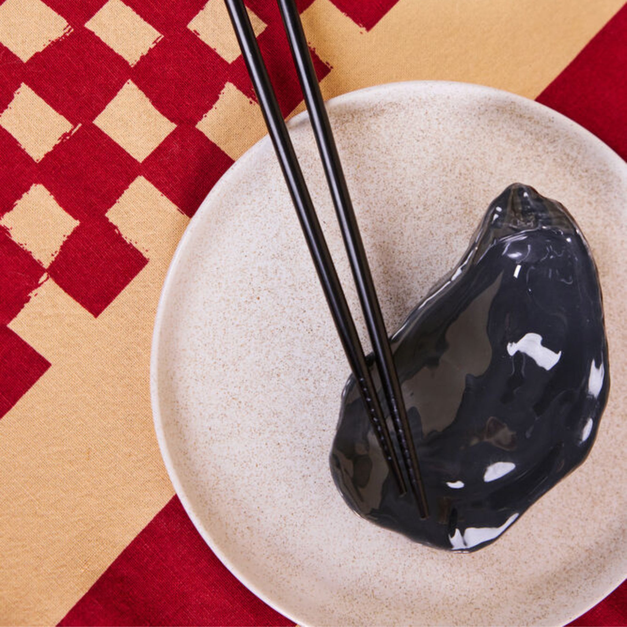 Een paar zwarte eetstokjes rust op een glanzend Byon Oester Schaaltje Grijs, gedekt op een beige bord op een rood en geel tafelkleed met patroon.