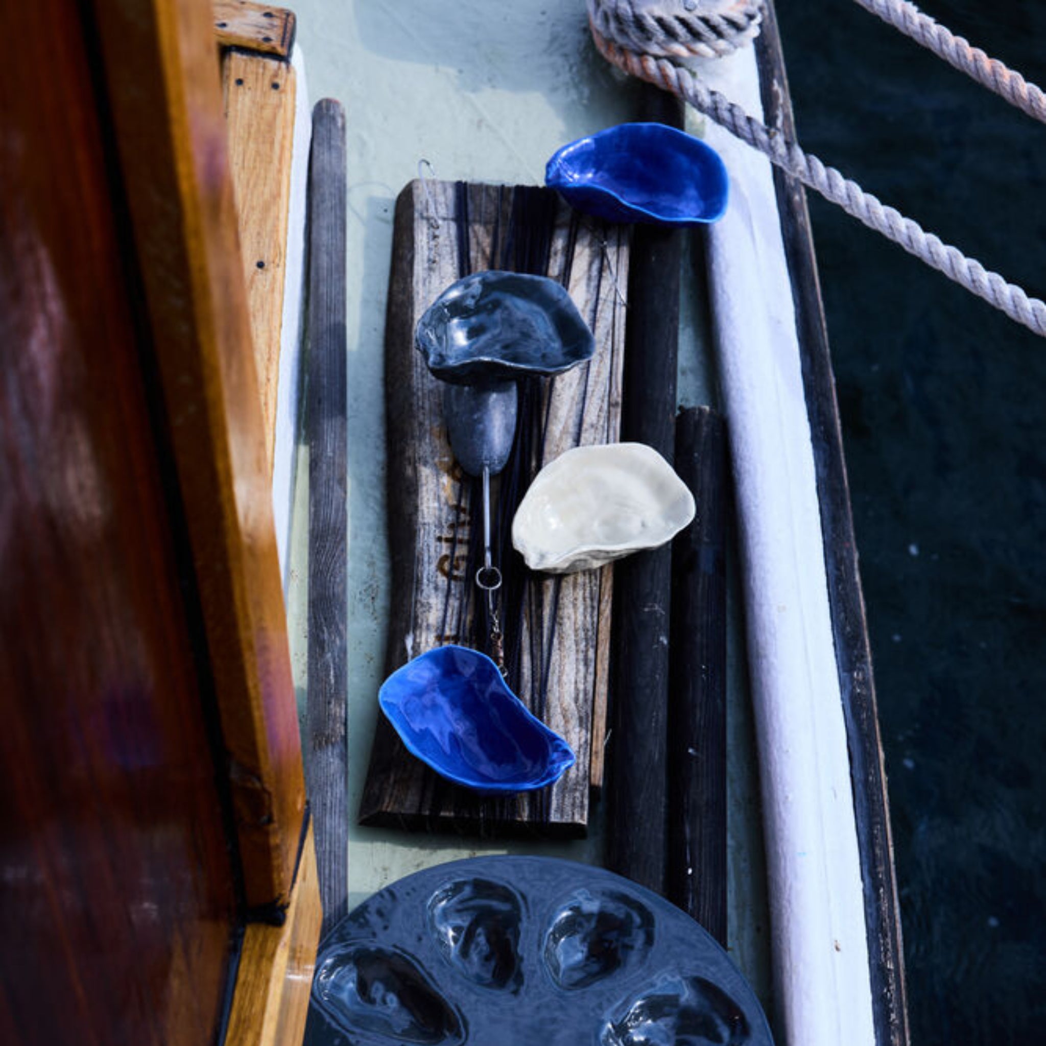 Byon Oester Schaaltje Wit keramische schaaltjes van Byon in blauw en wit, gerangschikt op een houten plank met een schelpdier notenkraker, uitgestald op een bootdek in de buurt van touwen en donker water.