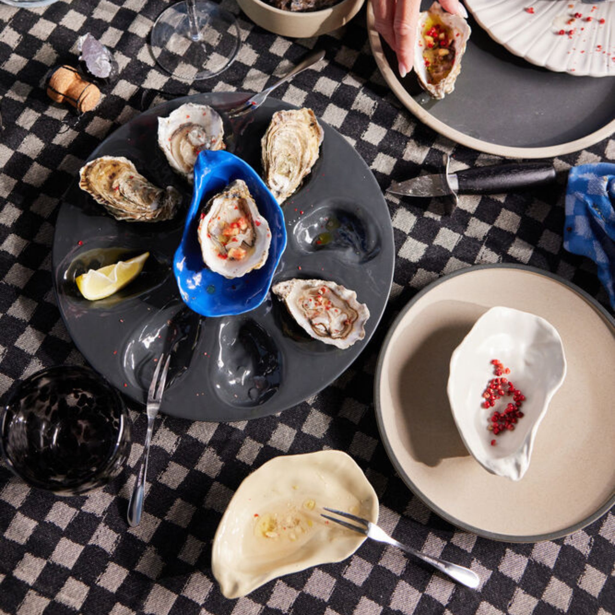 Een gedekte tafel met geopende oesters op een zwarte schaal, partjes citroen en borden, waaronder het Byon Oester Schaaltje Wit van Byon, op een geruit kleed. Een kreeft kraker rust vlakbij en roestvrijstalen keukengerei en glazen maken het tafereel compleet.