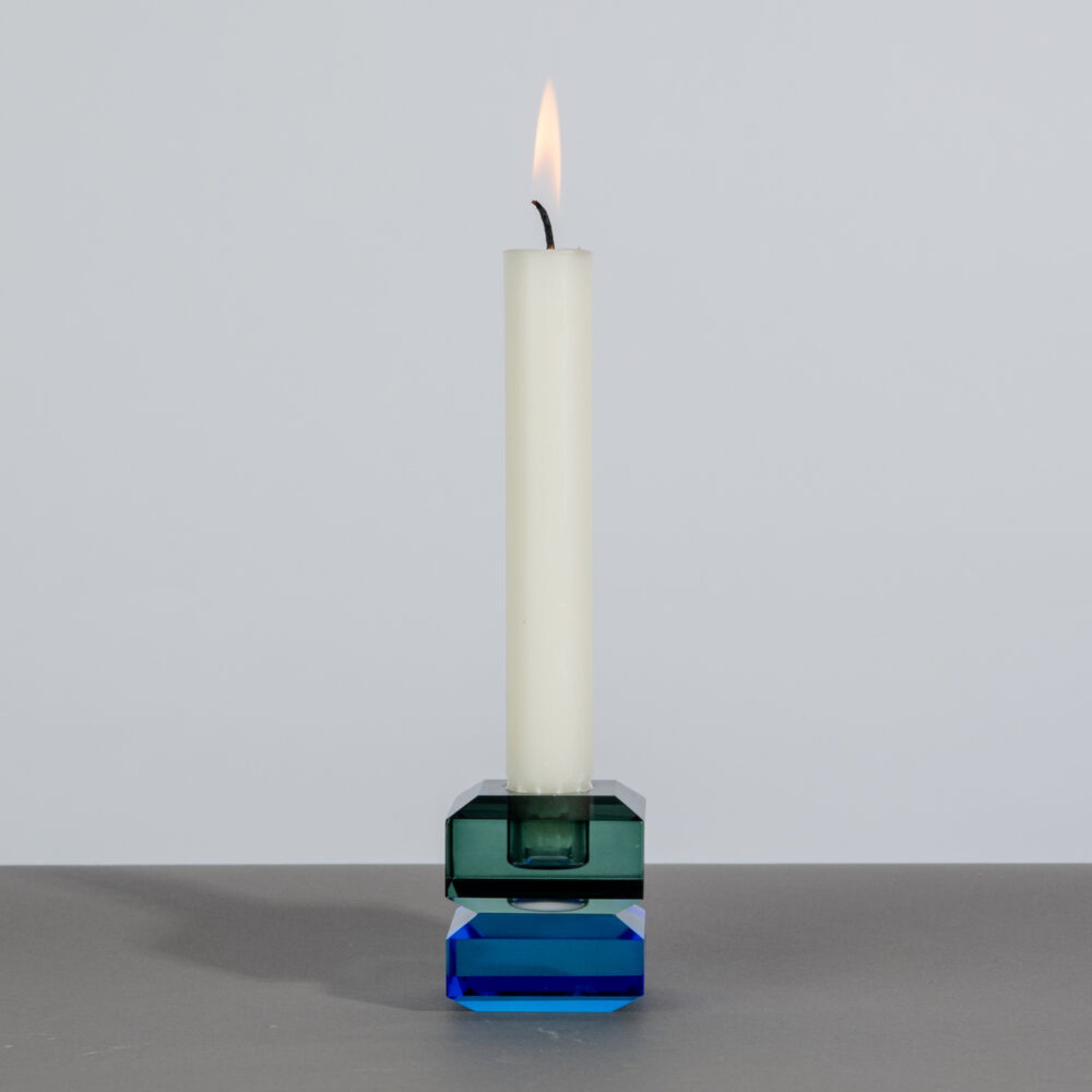 Een enkele witte kaars brandt in de Byon Ruby Kandelaar Groen/Blauw van Byon, gemaakt van doorschijnend blauw en groen kristalglas op een grijs oppervlak tegen een effen lichtgrijze achtergrond.