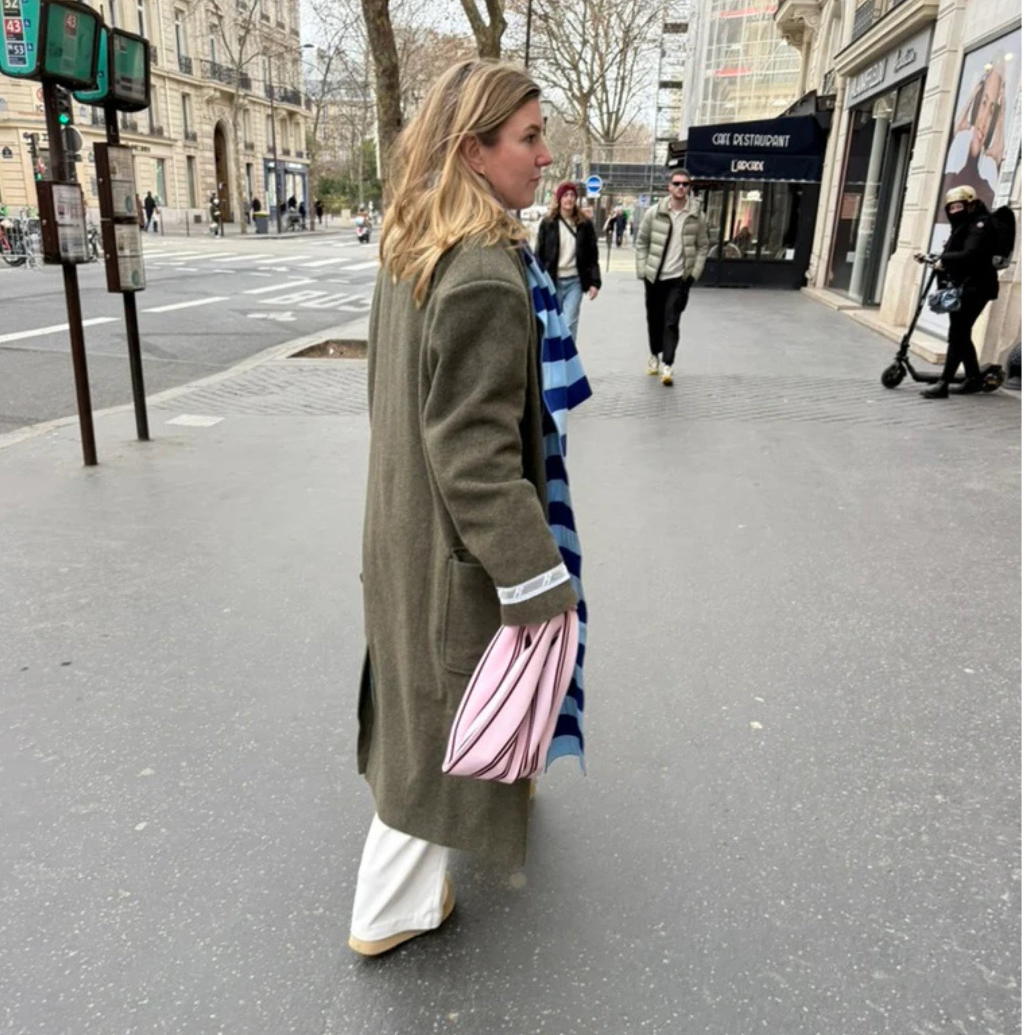Een vrouw in een lange groene jas, witte broek en beige schoenen loopt over een stadspad met de Caro Handtas Double Dash 2.0 Roland Larose van Caro Antwerp in de hand en draagt een blauw-wit gestreepte sjaal. Gebouwen en voetgangers zijn zichtbaar op de achtergrond.