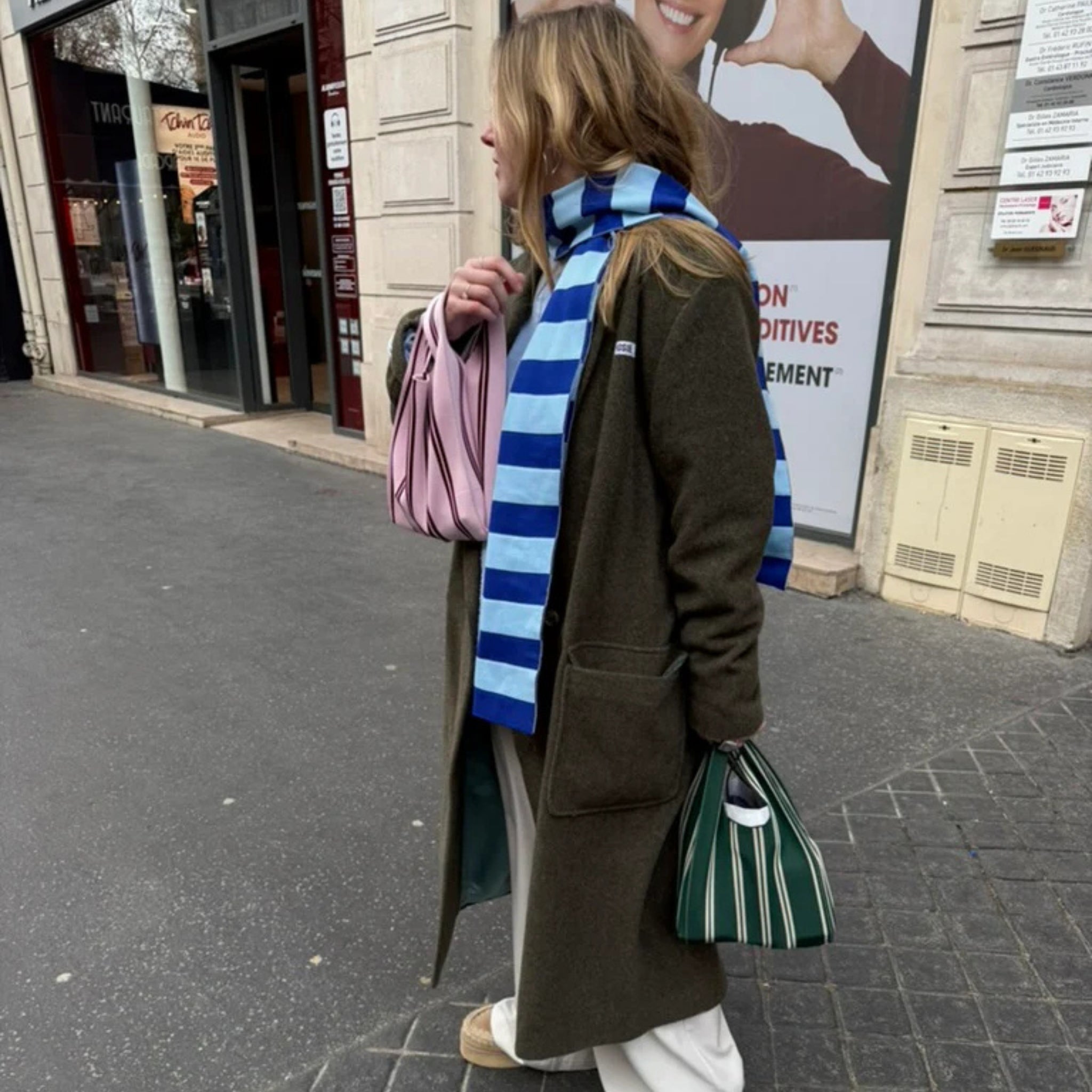 Een persoon in een lange donkere jas, blauwe en witte sjaal en witte broek staat op een trottoir in de stad met een roze tas en de Caro Antwerp Handtas Double Dash 2.0 Wimbledon - tijdloze kleuren. Hun haar zit los en ze kijken opzij.