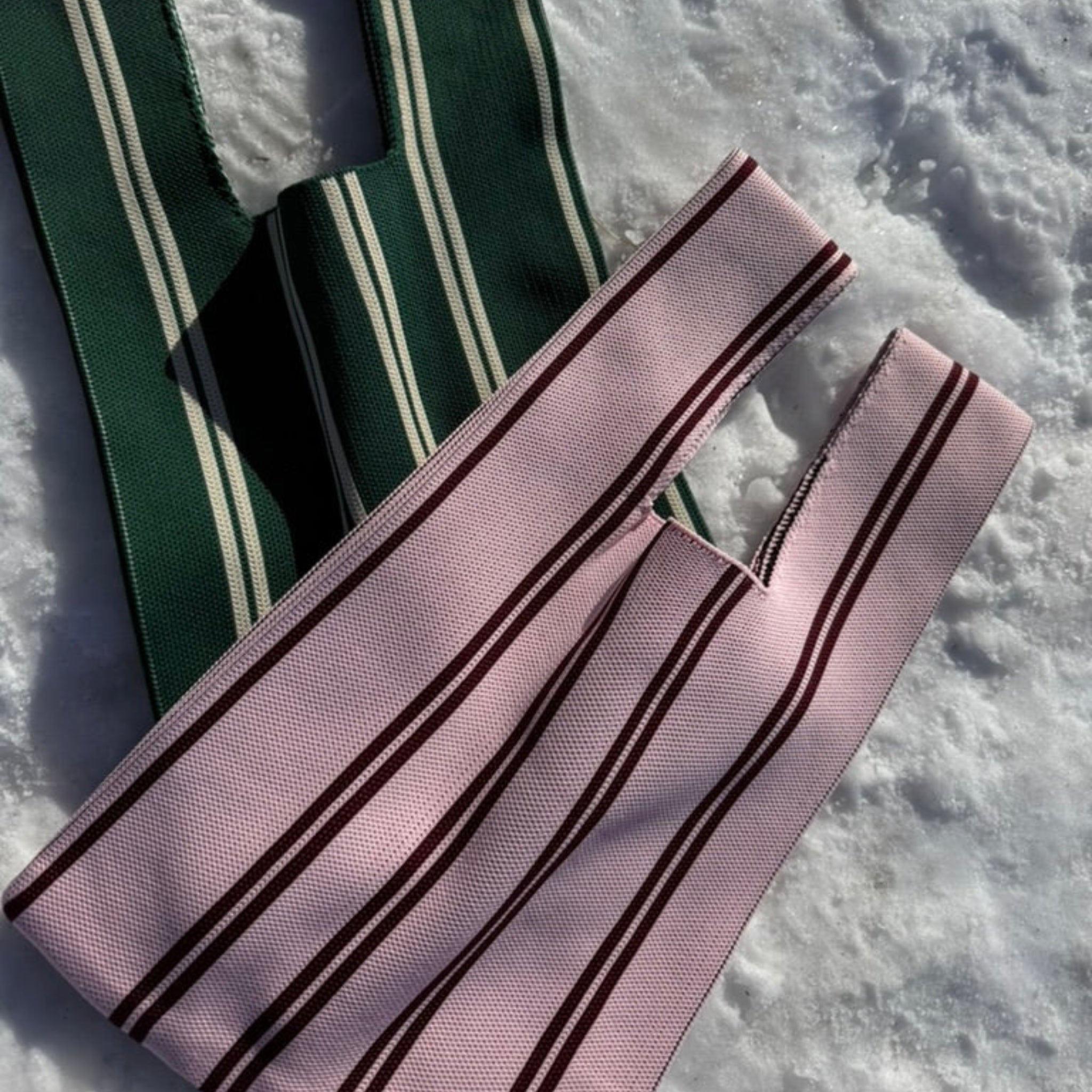 Twee Caro Antwerp Handtas Double Dash 2.0 Roland Larose tote bags-één groen/wit, één roze/bordeaux met verticale strepen-liggen op sneeuw in fel zonlicht. Deze stijlvolle geweven tassen van Caro Antwerp zijn perfect voor dagelijks gebruik.