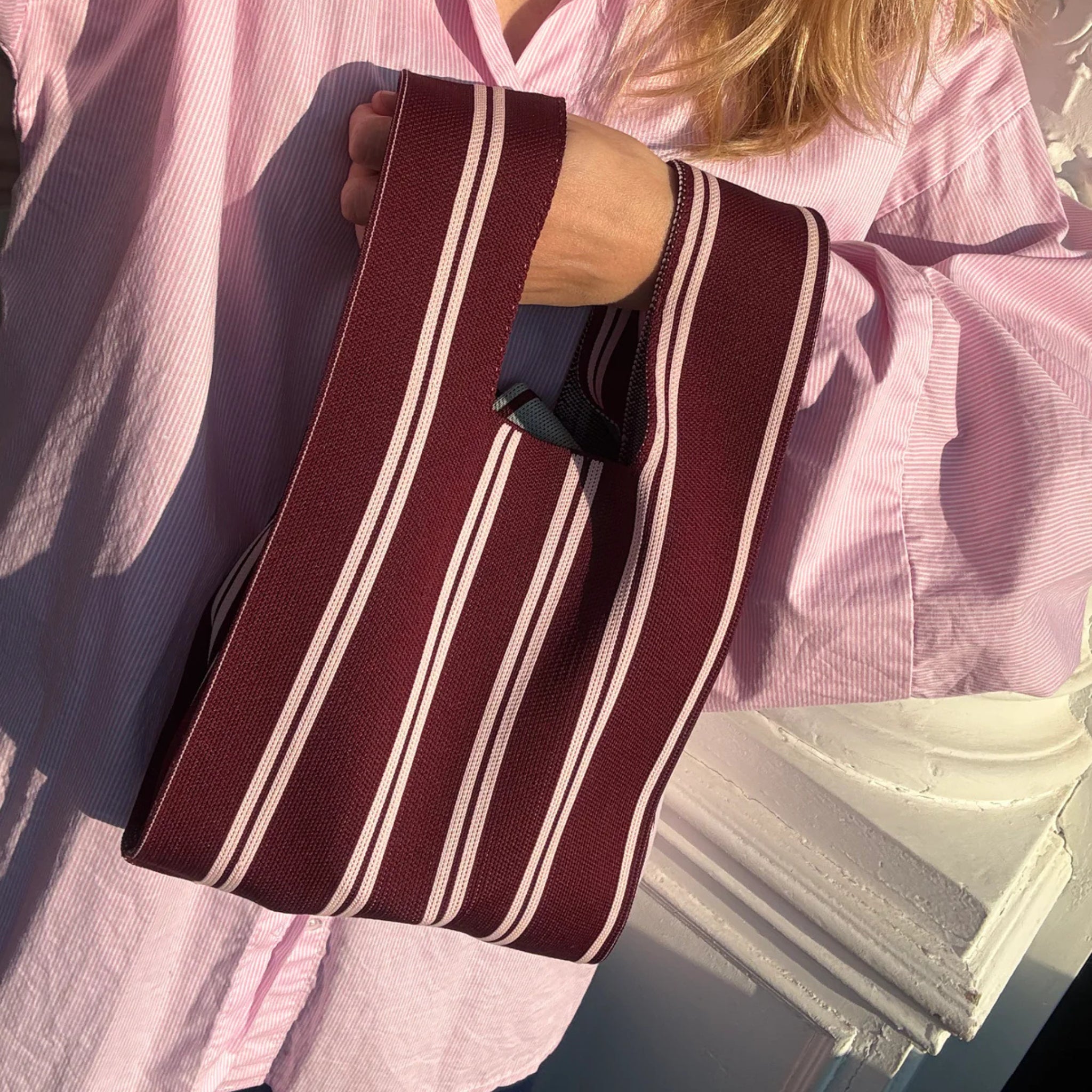 Een persoon in een lichtroze shirt houdt de Caro Handtas Double Dash Pink Promise van Caro Antwerp vast. Zonlicht werpt speelse schaduwen, die de vrolijke geest van het Pink Promise ontwerp weerspiegelen.