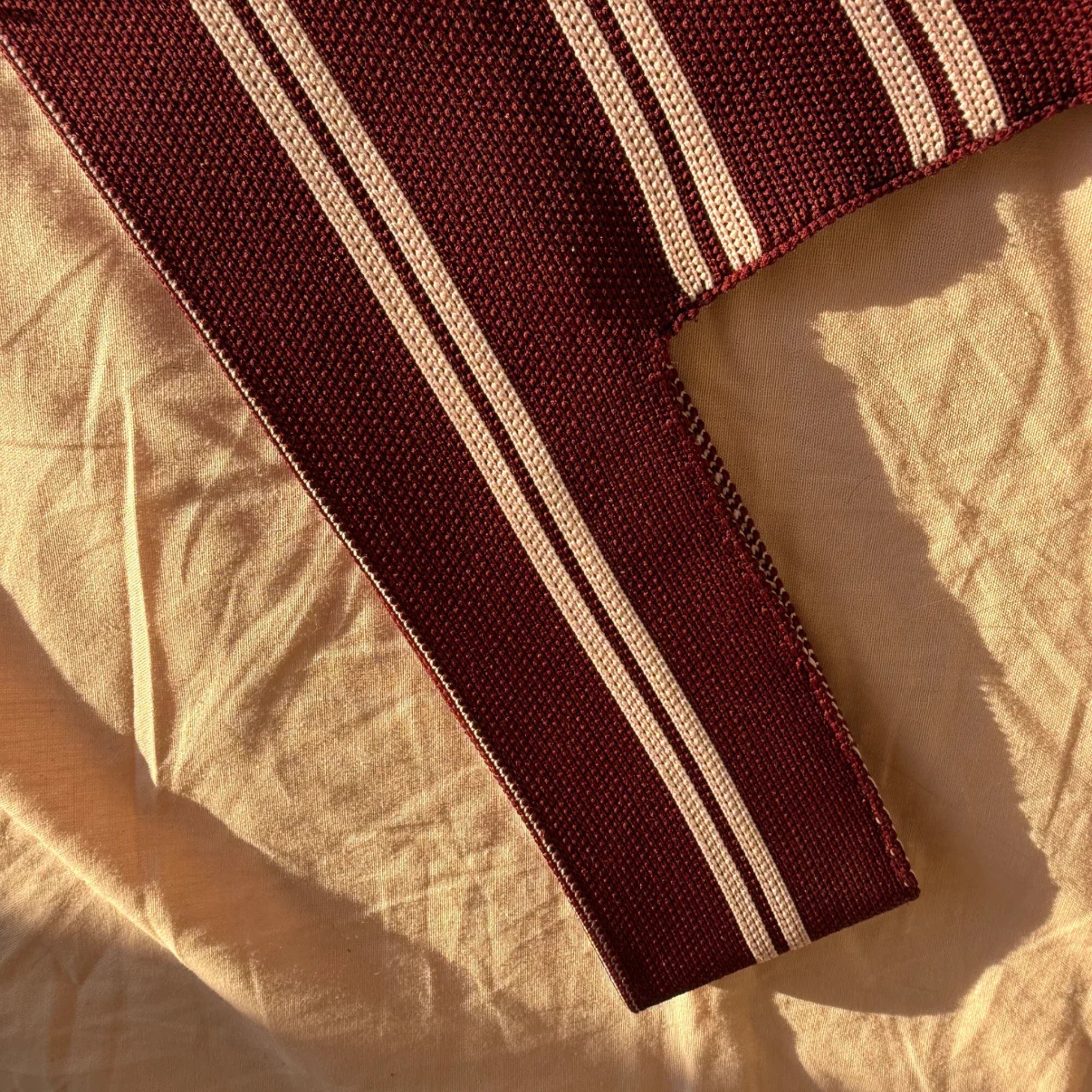 De Caro Antwerp Handtas Double Dash Pink Promise, met kastanjebruine stof met twee witte strepen, ligt op een verfrommeld beige doek en baadt in warm zonlicht.