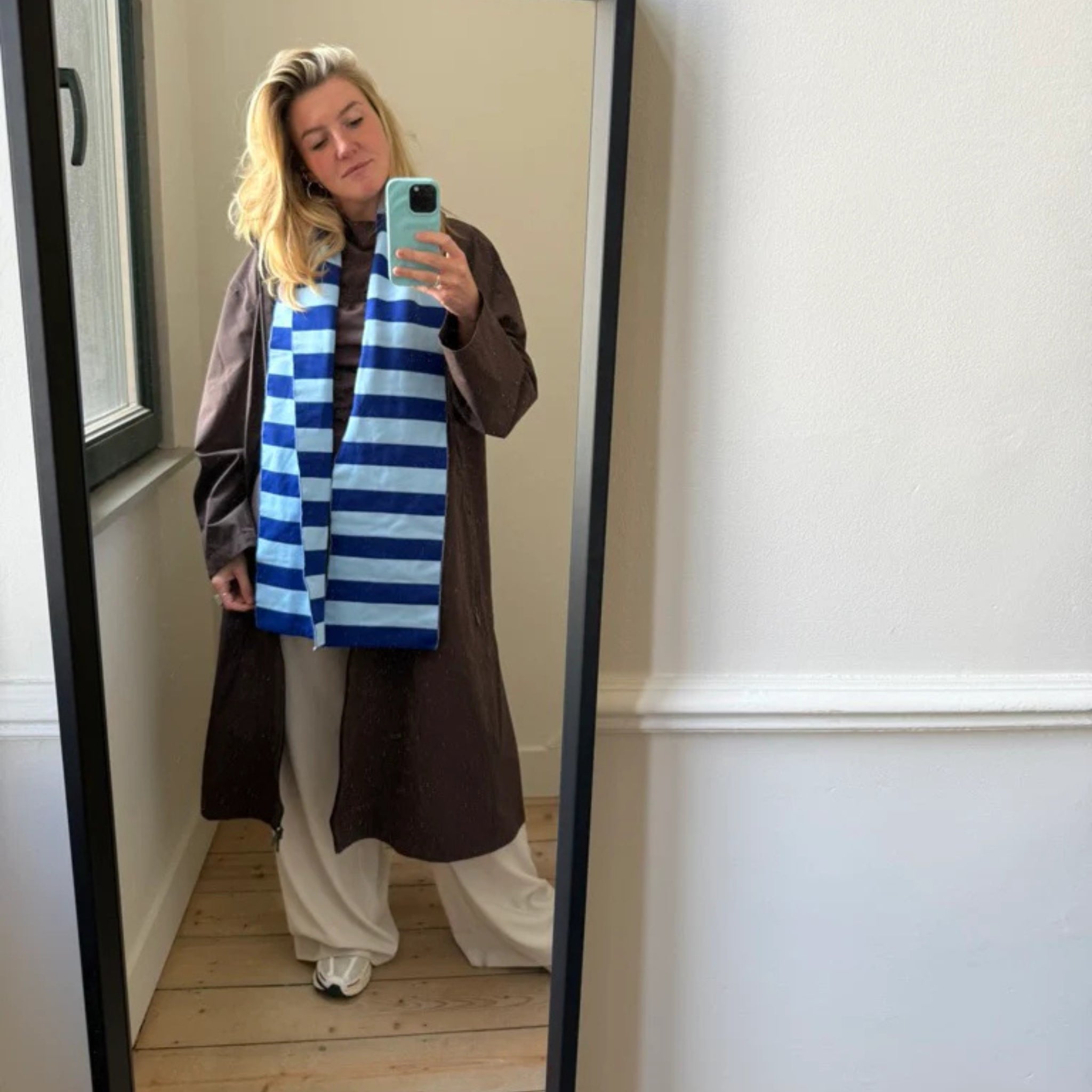 Een persoon neemt een selfie voor een spiegel en draagt een lange bruine jas, een losse witte broek, sneakers en de Caro Antwerp Caro Sjaal Ace Ice Ice Baby sjaal. De kamer heeft lichte muren en houten vloeren.